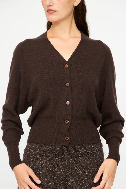 A woman wears the Cordera Merino and Cashmere Cardigan in brown, featuring brown buttons, paired with textured dark pants, standing against a plain white background.