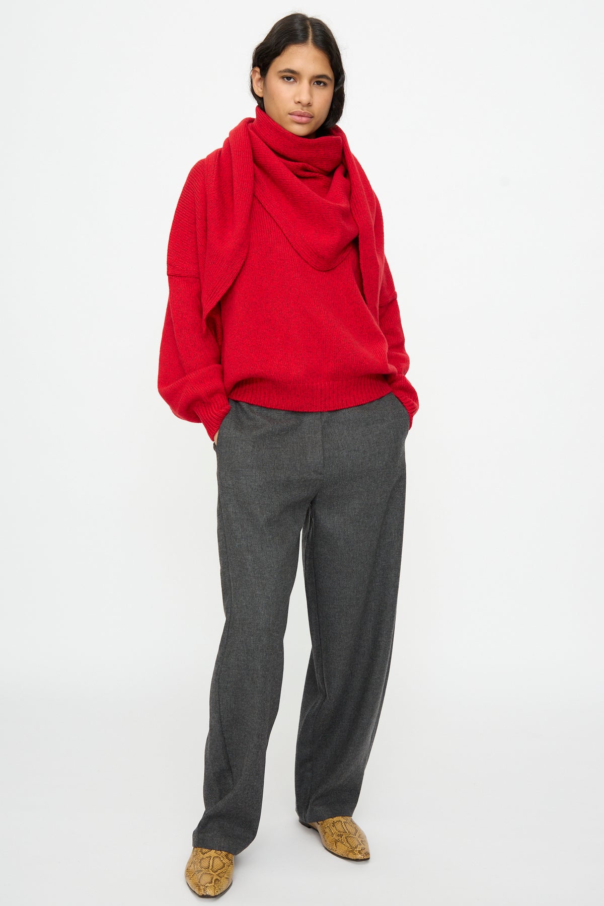 A person wearing a bright red sweater and a Cordera Merino and Cashmere Maxi Bandana in Red draped around their neck, paired with grey trousers and patterned ankle boots.