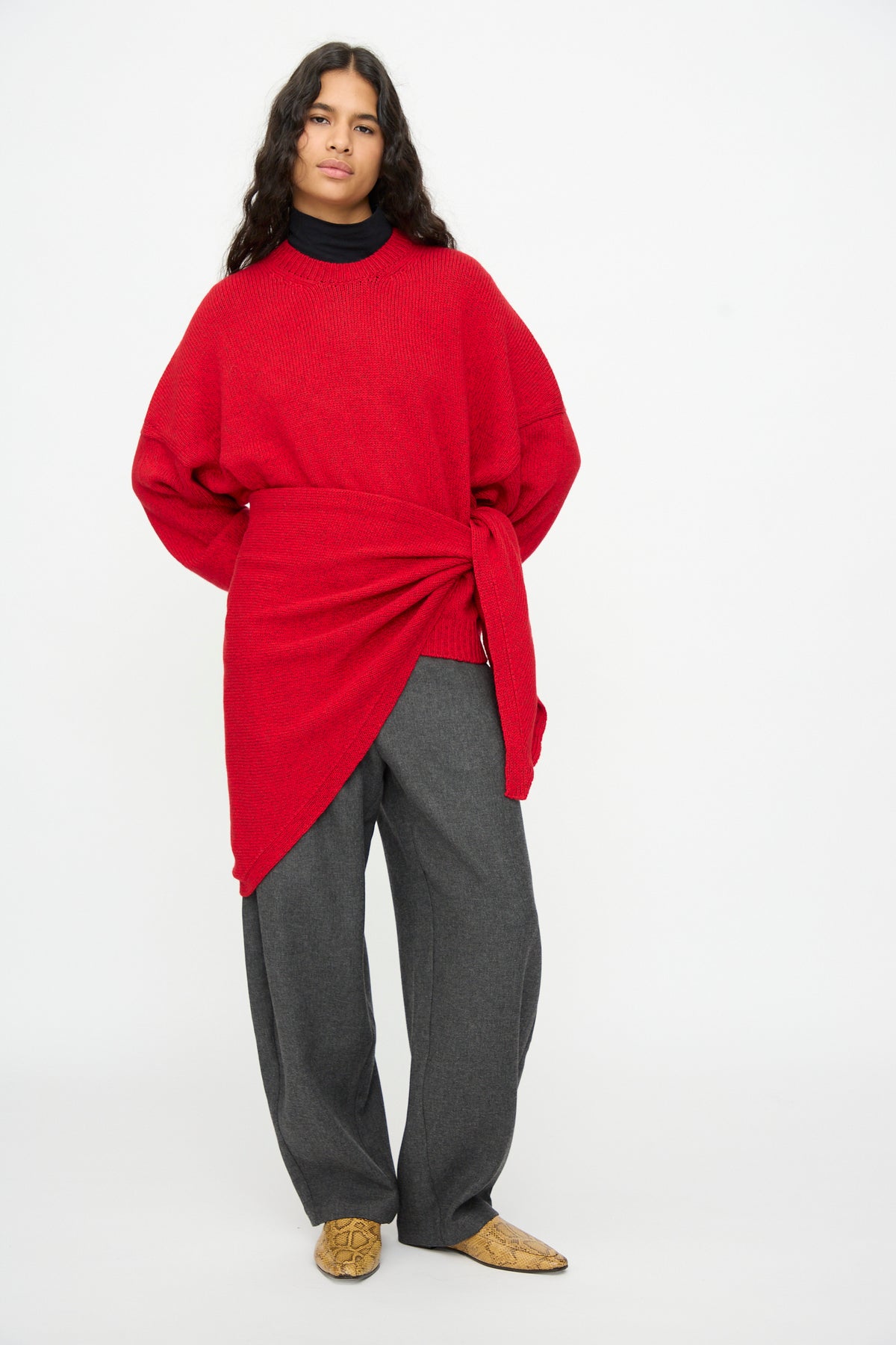A person wears a red Merino and Cashmere Maxi Bandana with an asymmetrical wrapped waist tie over a black top and grey trousers, paired with snakeskin-print ankle boots.