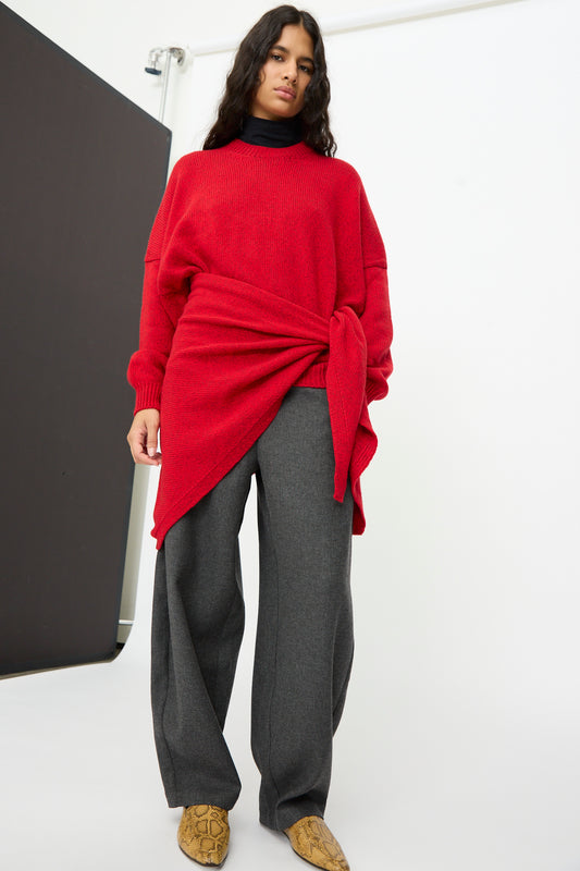 A person wearing a Cordera Merino and Cashmere Maxi Bandana in Red styled as a long-sleeved triangular top at the waist, with dark grey trousers and patterned ankle boots.