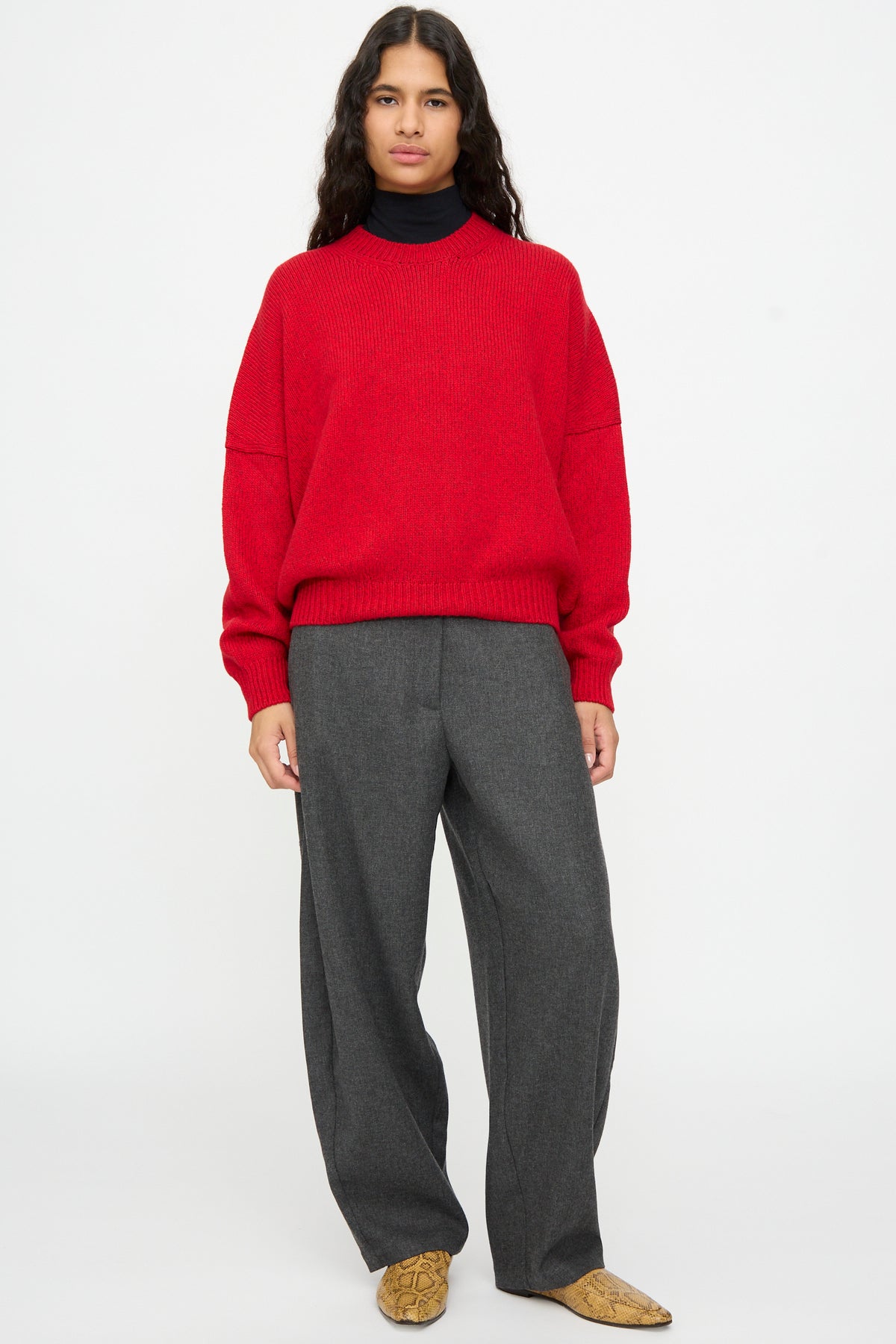 A person wears a Cordera Merino and Cashmere Sweater in Red over a dark turtleneck, paired with gray trousers and patterned pointed-toe shoes.