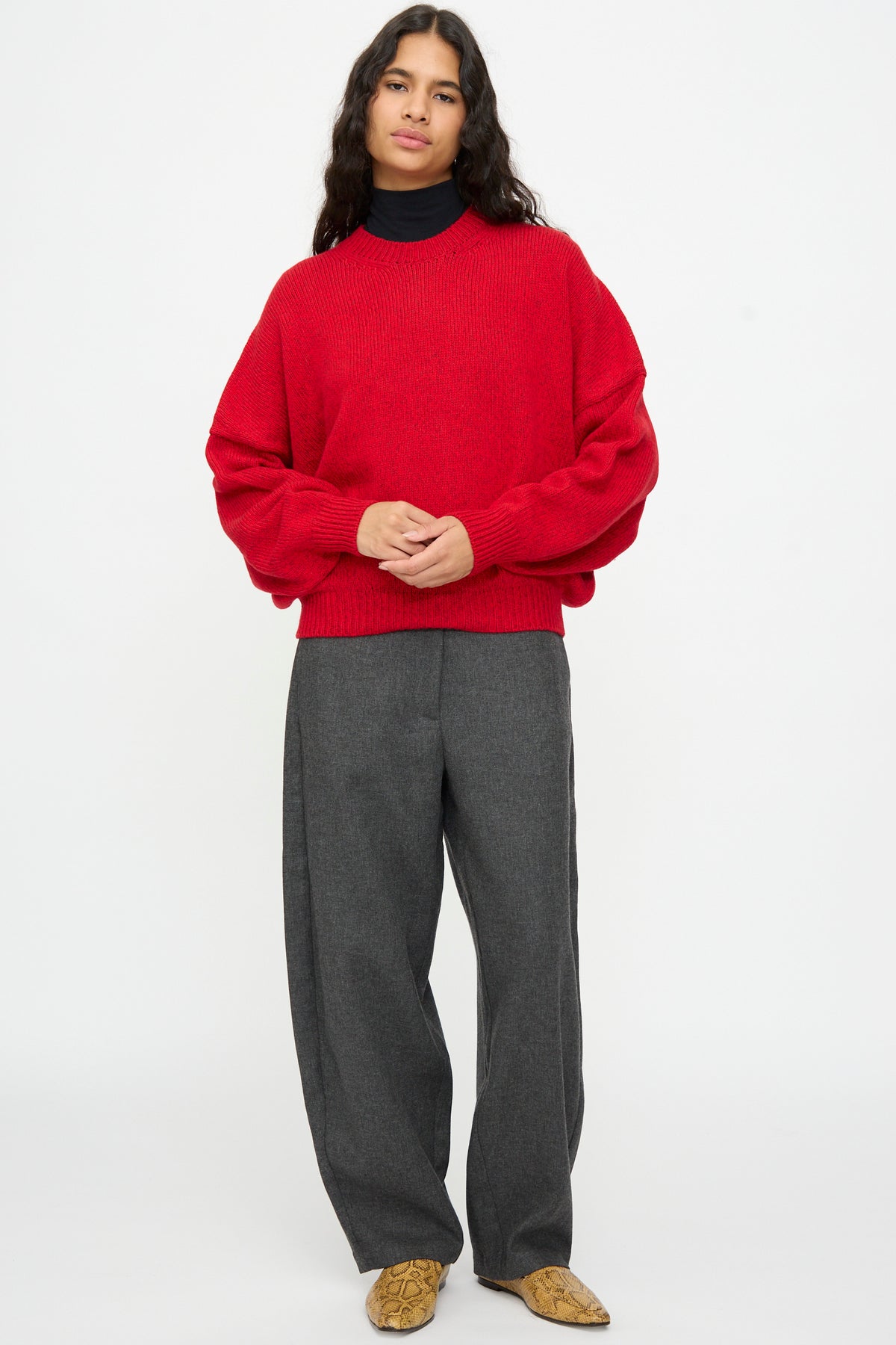 Person wearing an oversized, batwing-sleeve Merino and Cashmere Sweater in Red over a black turtleneck, paired with charcoal grey trousers and snakeskin-patterned loafers.