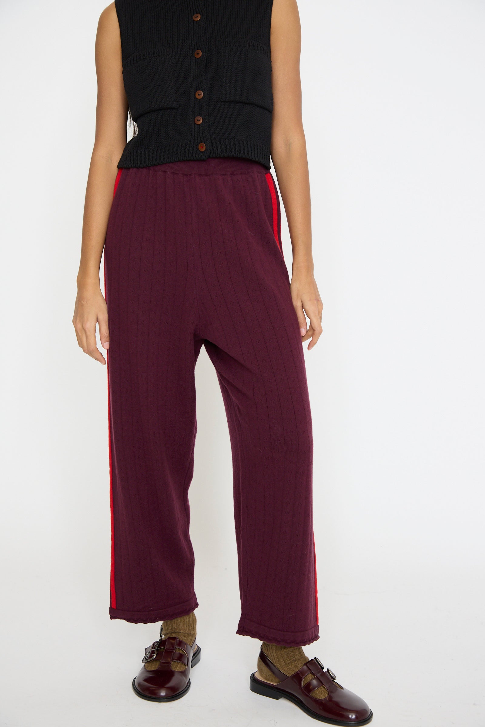 A person is dressed in a black sleeveless top and the Cordera Merino Perlé Pant in Burgundy, featuring a red stripe. 