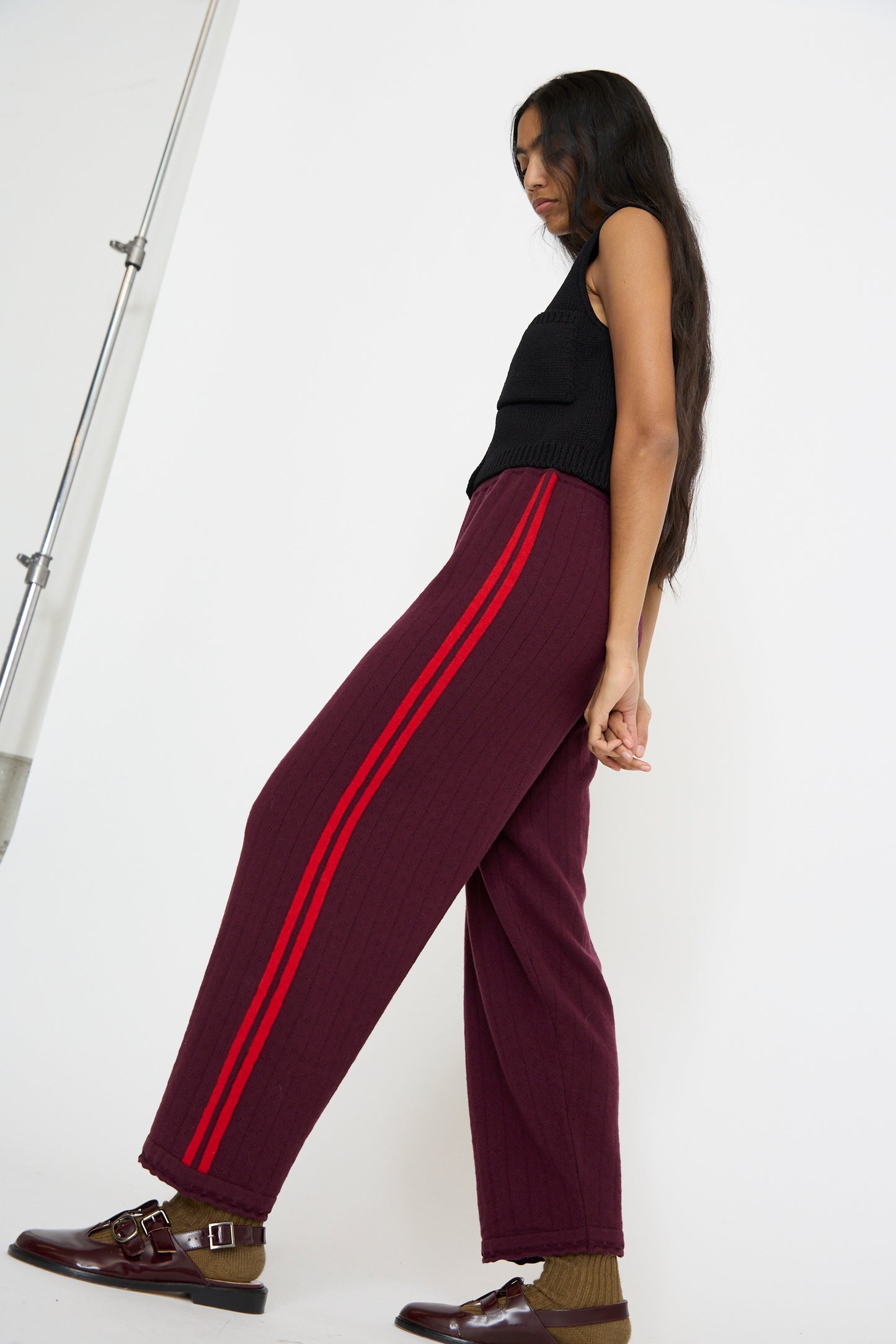 A person with long hair stands casually, wearing a black sleeveless top, Cordera's Merino Perlé Pant in burgundy featuring red stripes, and brown shoes.