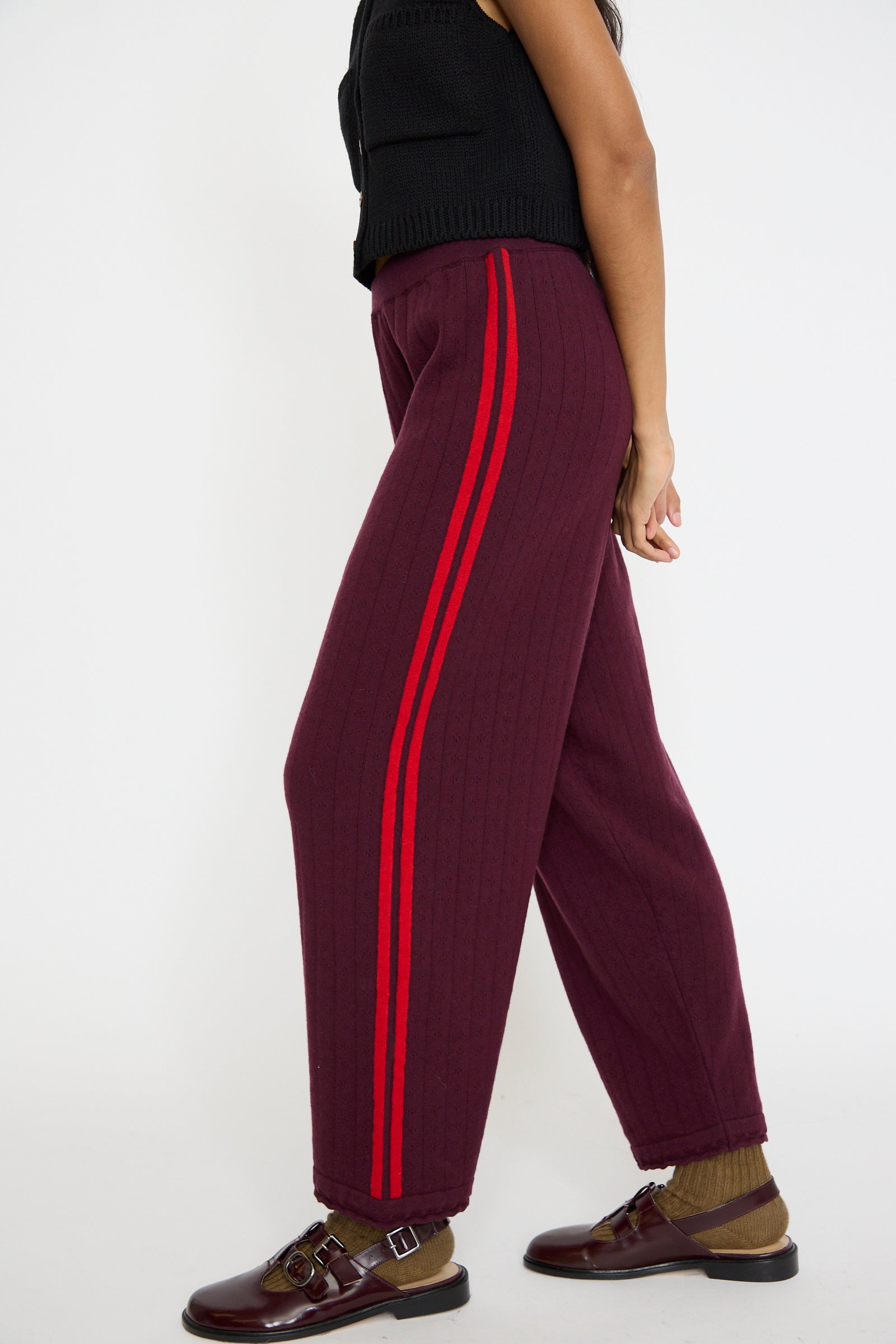 A person is standing sideways wearing Cordera's Merino Perlé Pant in Burgundy with red side stripes, paired with a black top, and complemented by brown socks and shoes.