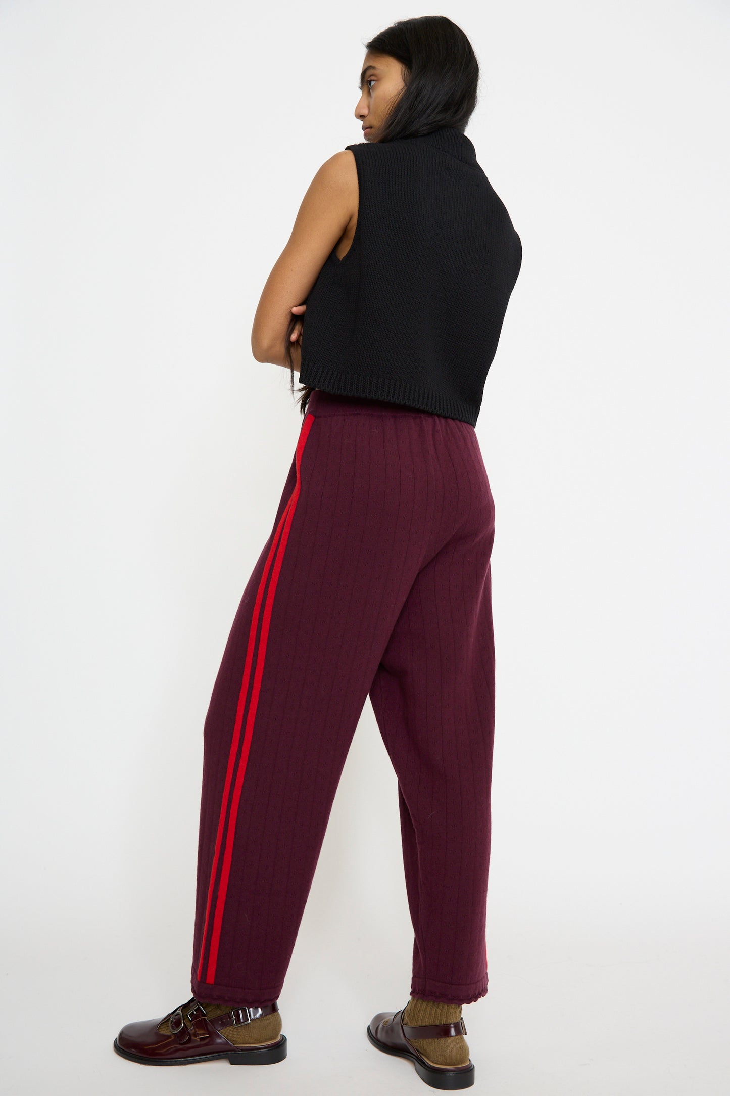 A person is standing sideways against a plain white background, wearing the Merino Perlé Pant in Burgundy by Cordera and a sleeveless black sweater, featuring relaxed fit pants with red stripes.