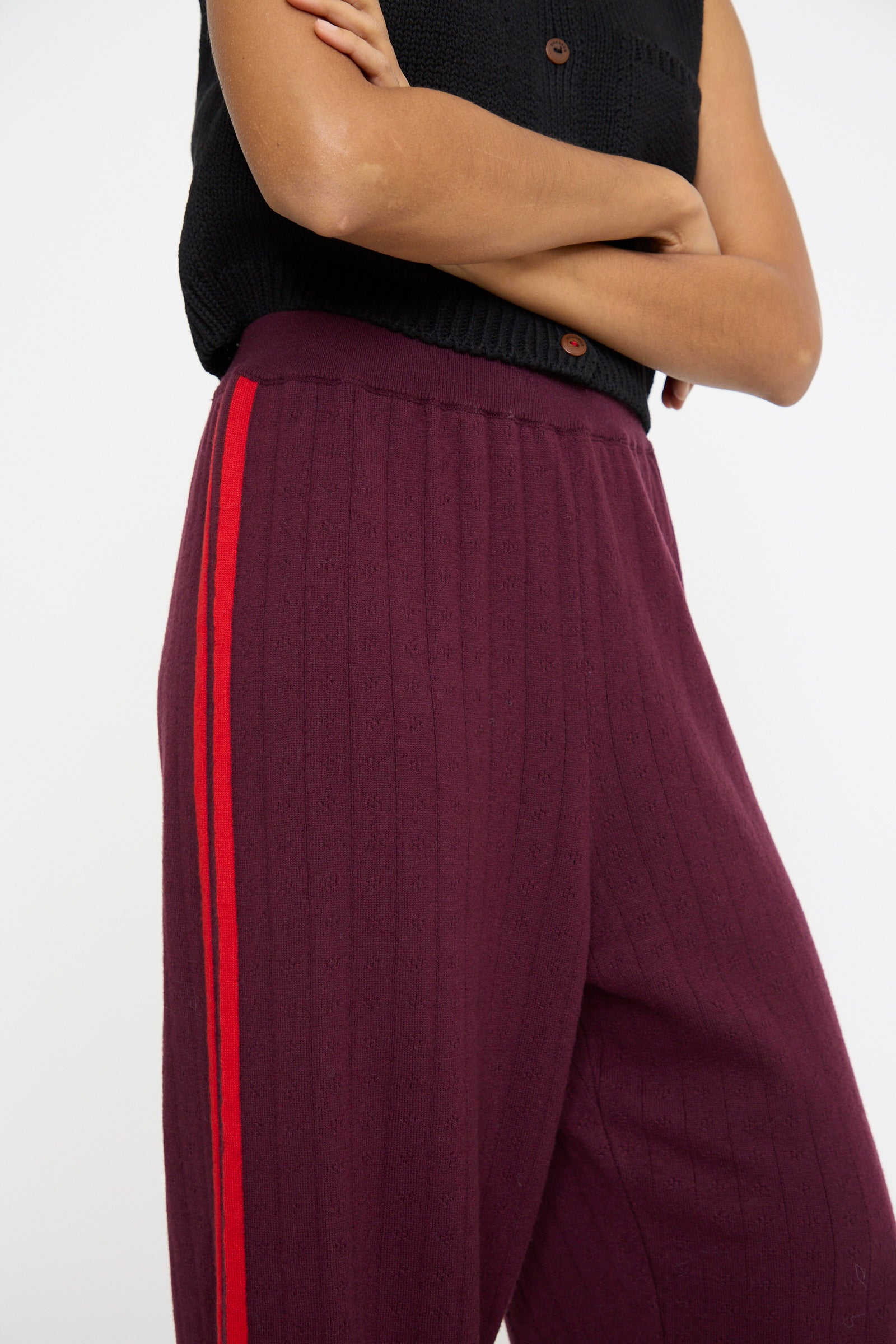 A person in Cordera's sleeveless black top and Merino Perlé Pant in burgundy with a red stripe, arms crossed against a plain backdrop. 