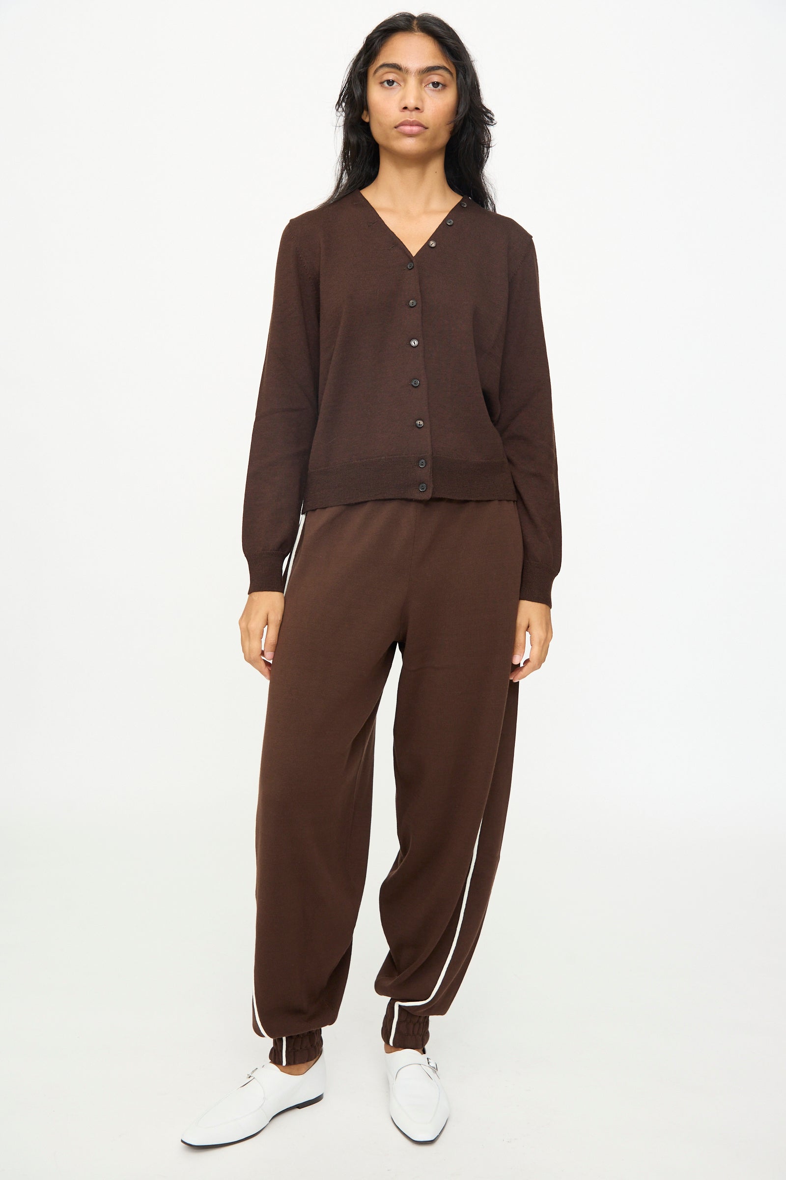 A person stands facing forward against a plain white background, wearing the Cordera Merino Wool Button Cardigan in Brown paired with matching pants featuring a white stripe near the ankles, and white shoes.