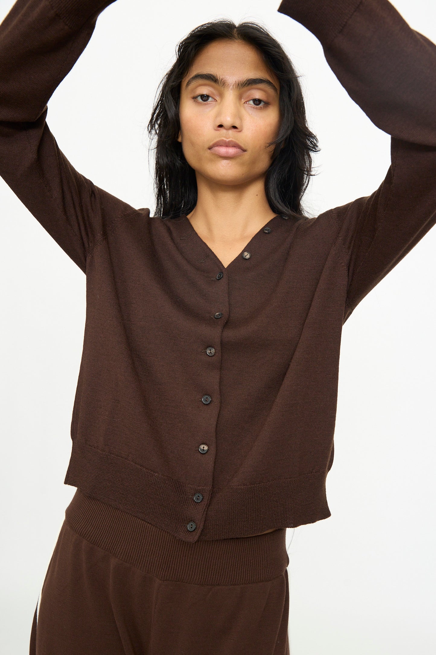 A person wearing the Cordera Merino Wool Button Cardigan in Brown with matching brown pants, standing against a plain white background with arms raised overhead.