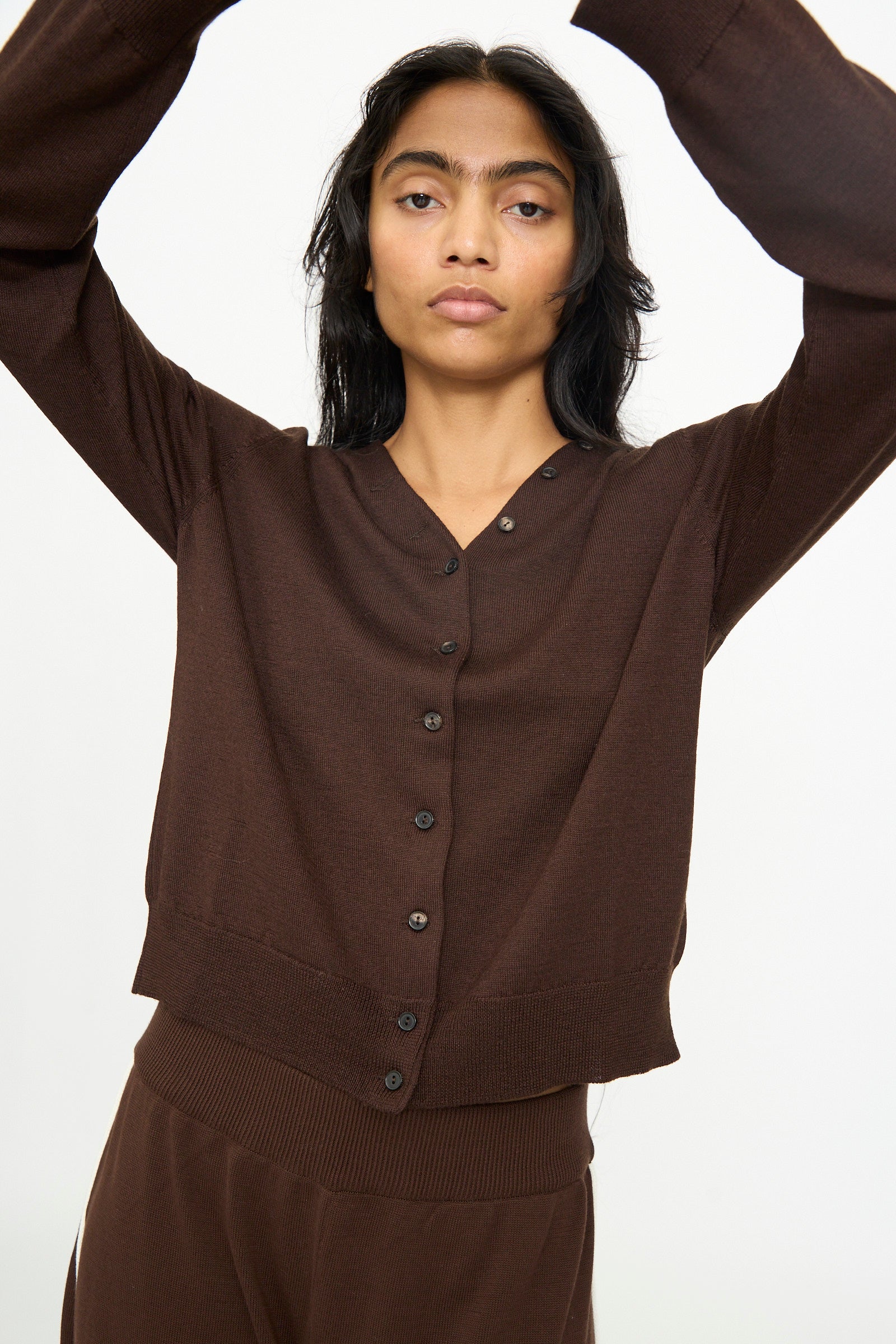 A person wearing the Cordera Merino Wool Button Cardigan in Brown with matching brown pants, standing against a plain white background with arms raised overhead.