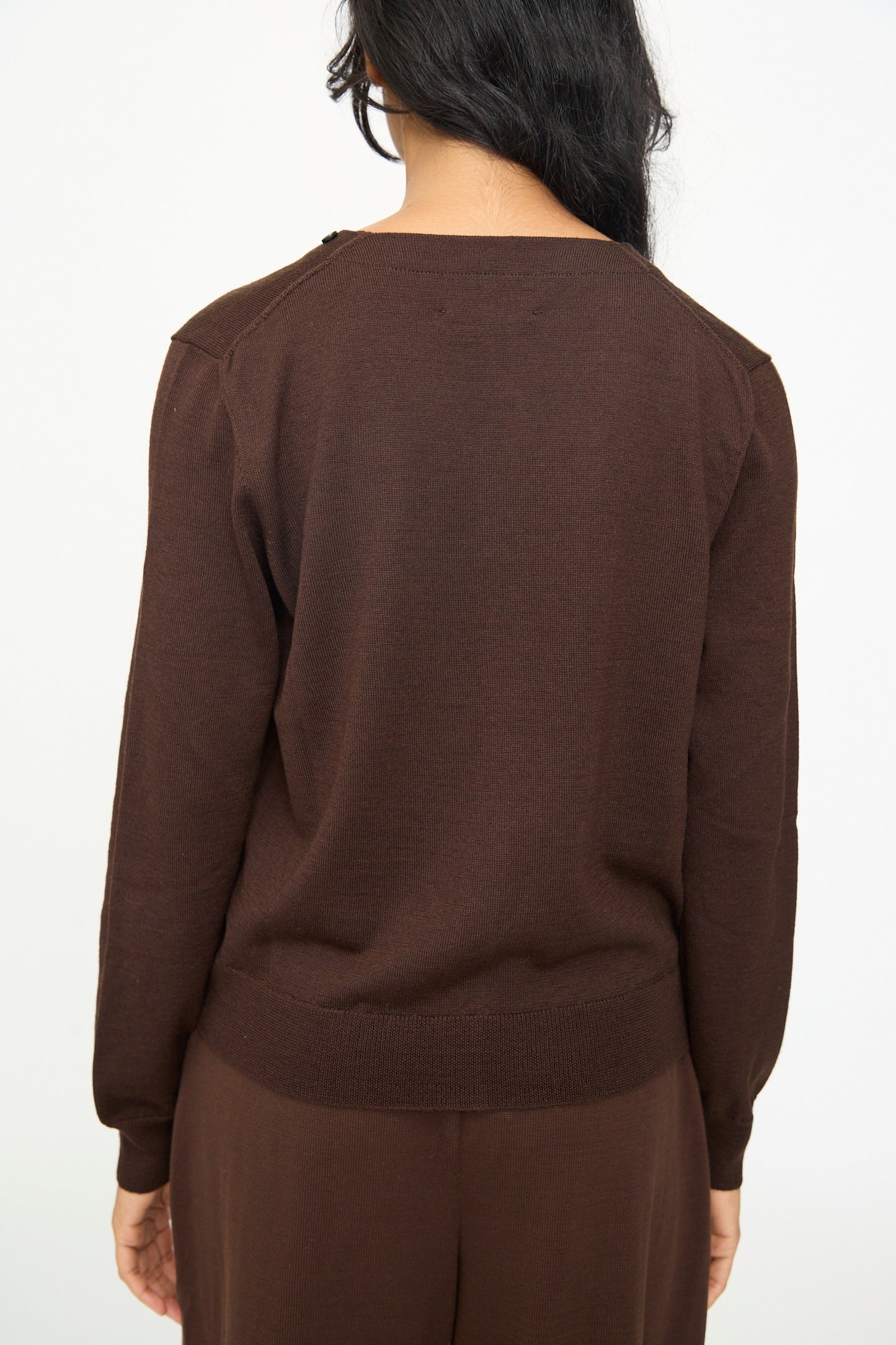 A person with long dark hair is shown from the back against a plain white background, wearing the Cordera Merino Wool Button Cardigan in Brown paired with matching brown pants.