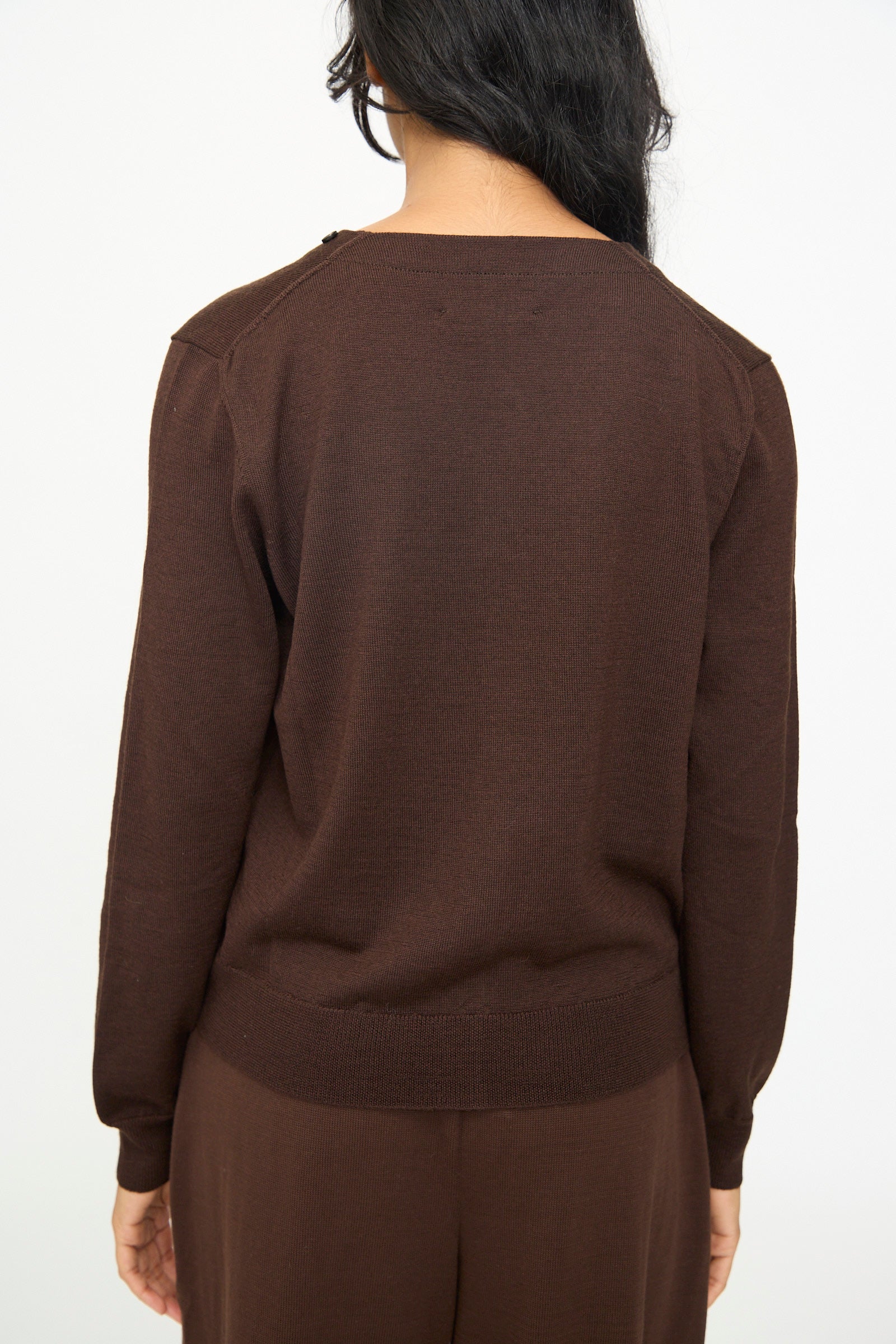 A person with long dark hair is shown from the back against a plain white background, wearing the Cordera Merino Wool Button Cardigan in Brown paired with matching brown pants.