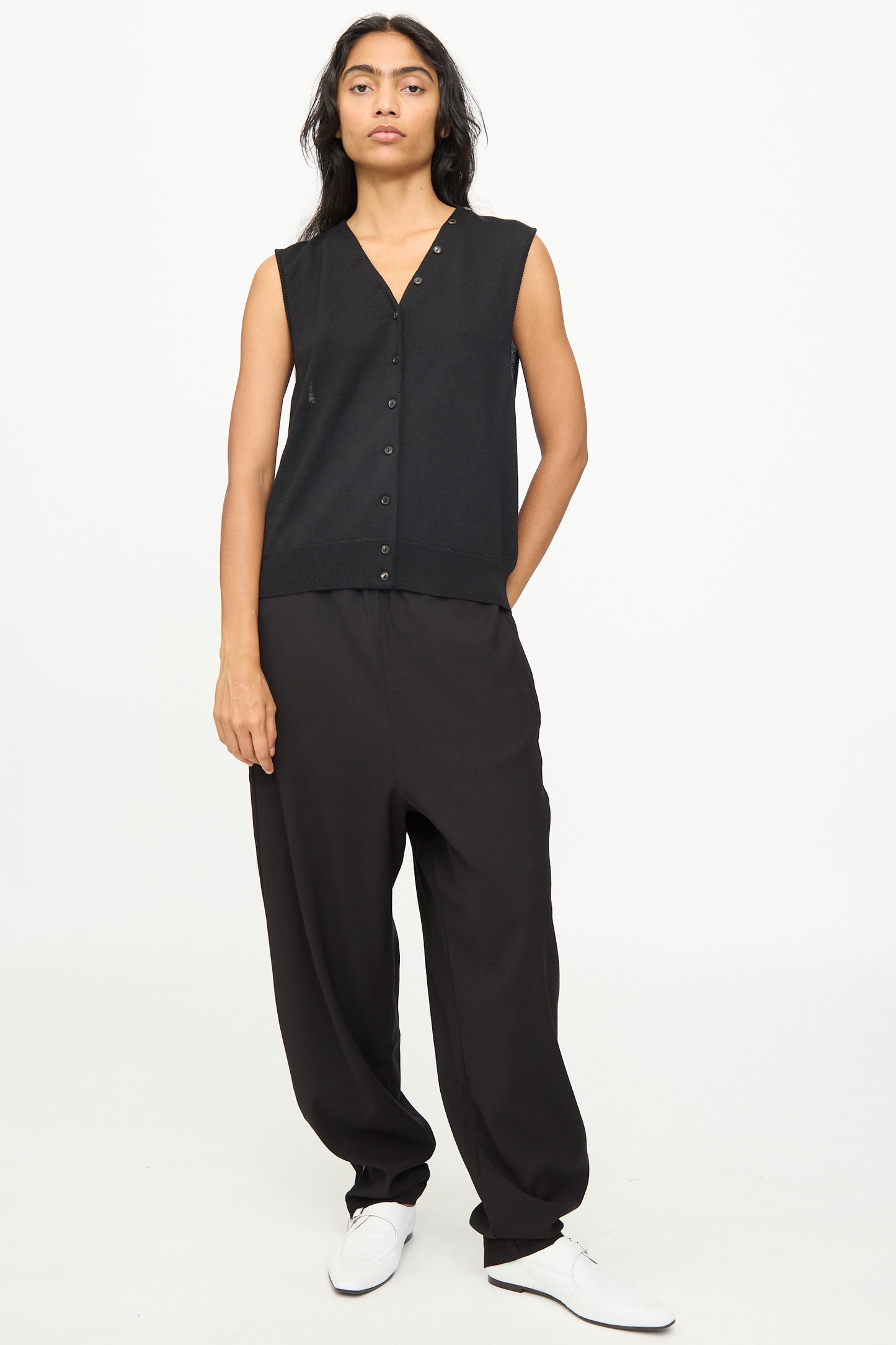 A person stands against a white background, wearing Cordera’s Merino Wool Button Waistcoat in Black—crafted in Spain—paired with loose black pants and white shoes, displaying a neutral expression and relaxed posture.