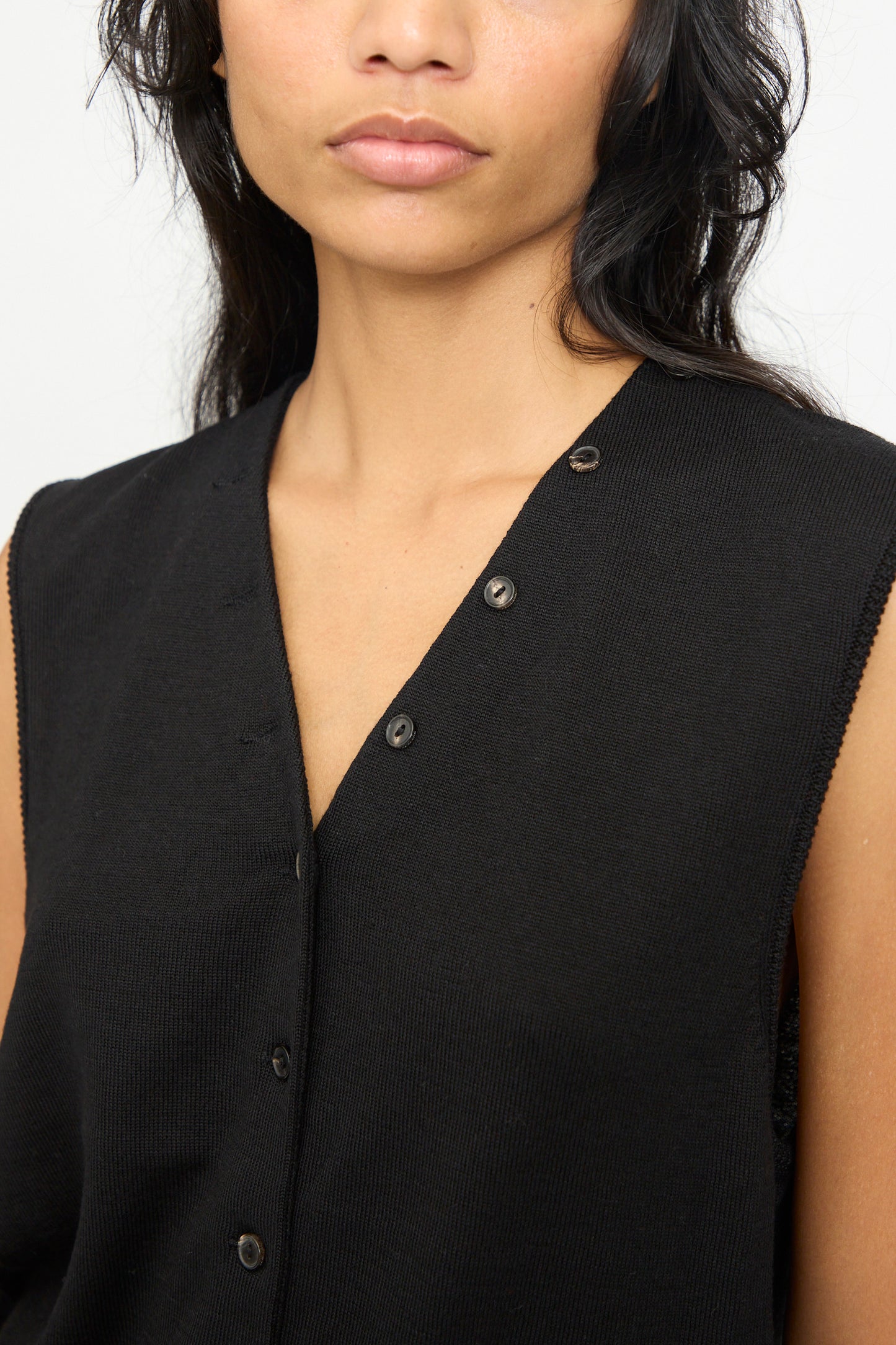 A woman models the Cordera Merino Wool Button Waistcoat in Black—a sleeveless, button-up top featuring visible black buttons—photographed from the shoulders to just above the lips against a plain background.