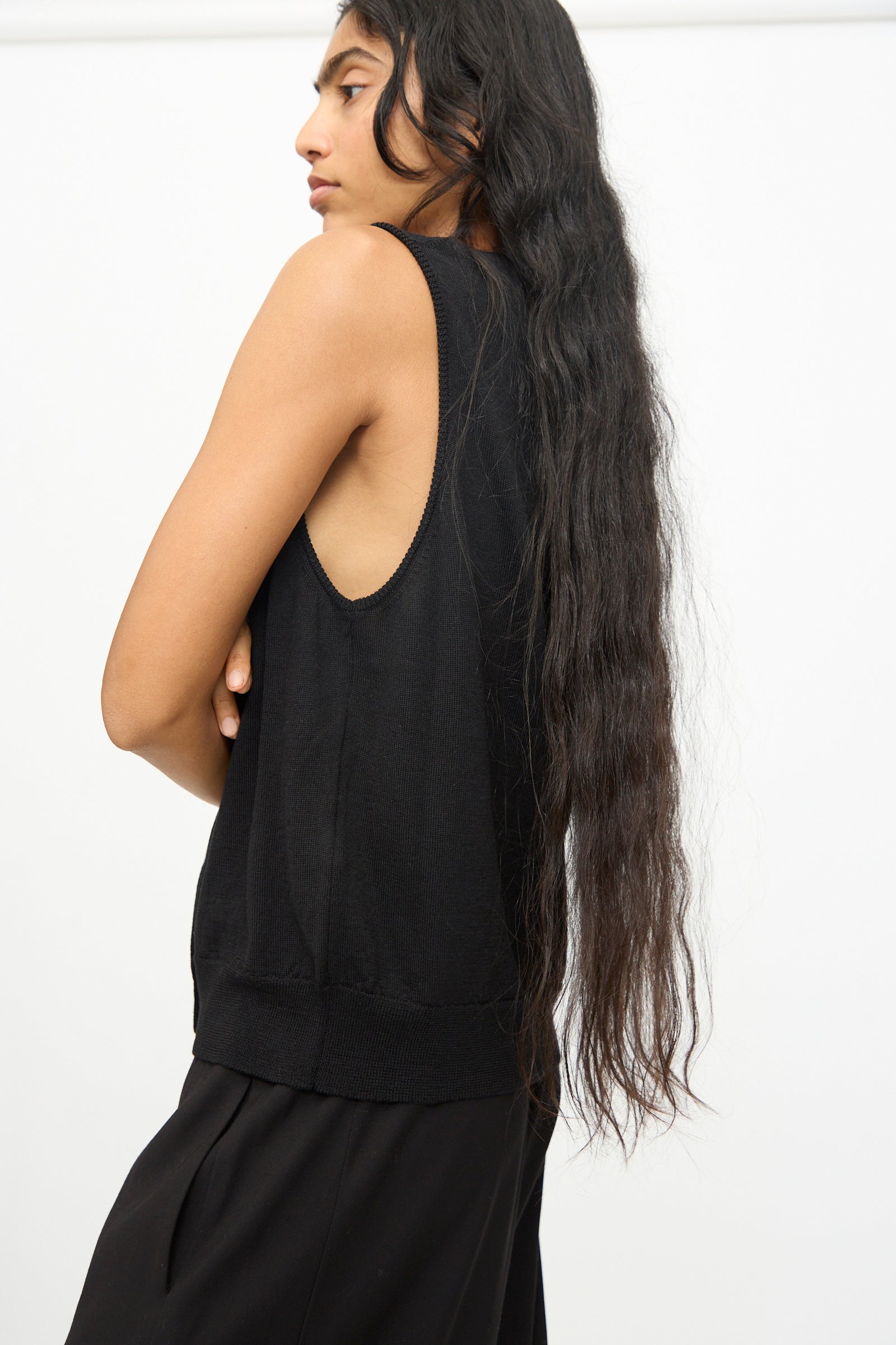 A person with long dark hair stands sideways against a plain white background, arms crossed, wearing Cordera's Merino Wool Button Waistcoat in Black paired with black pants.