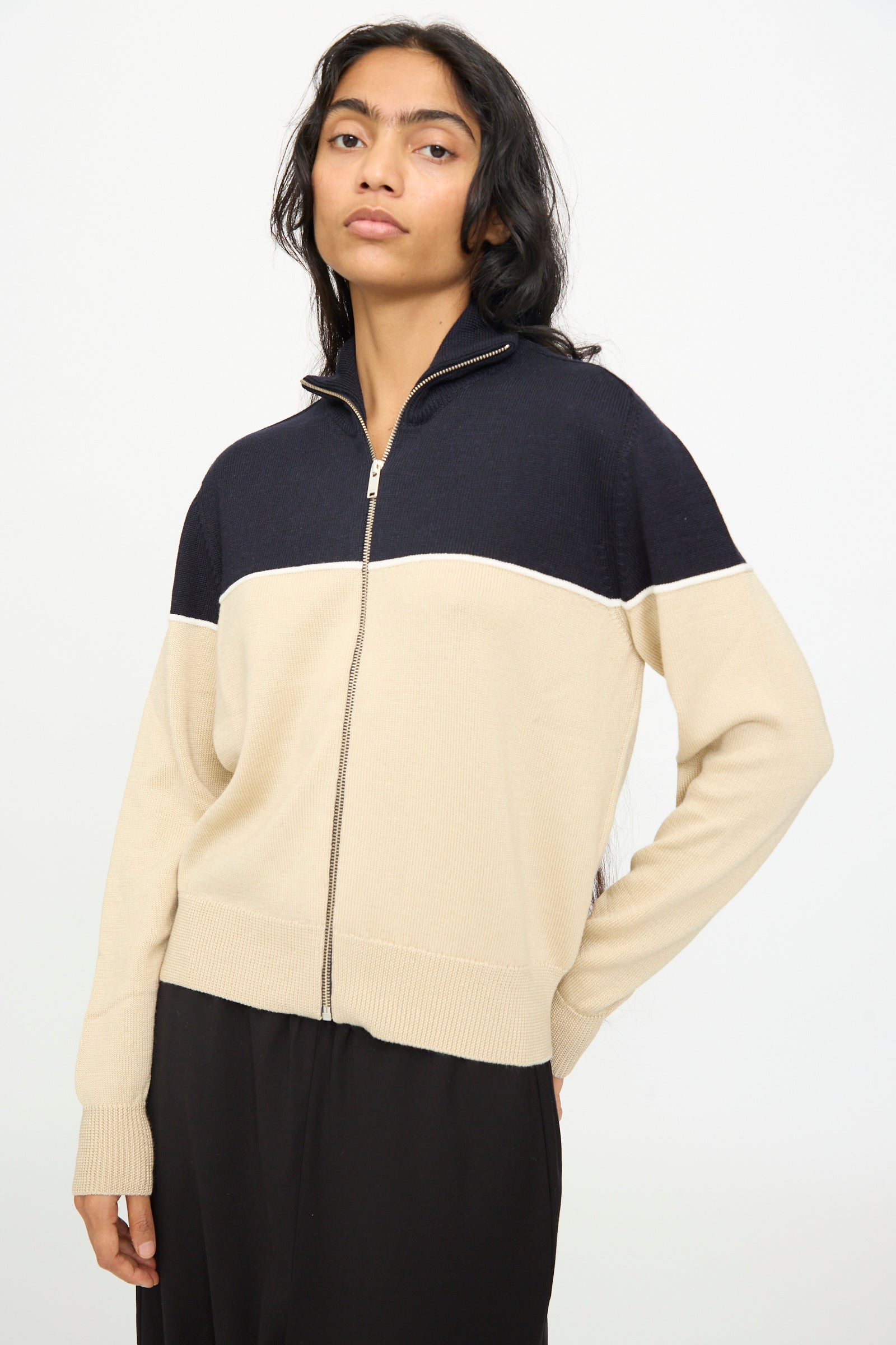 A woman stands against a plain white background wearing Cordera's Merino Wool Cardigan in Colorblock—crafted in Spain from extrafine merino wool, featuring beige and navy colorblocking, long sleeves, and paired with black pants.