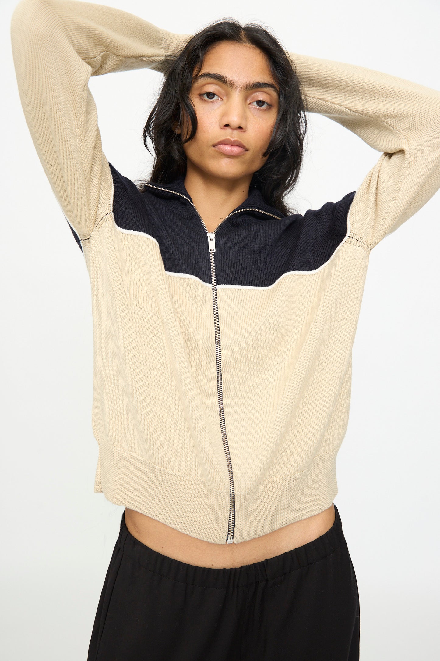 A person stands against a plain light background with arms raised behind their head, wearing the Cordera Merino Wool Cardigan in Colorblock, an extrafine merino wool piece expertly made in Spain.