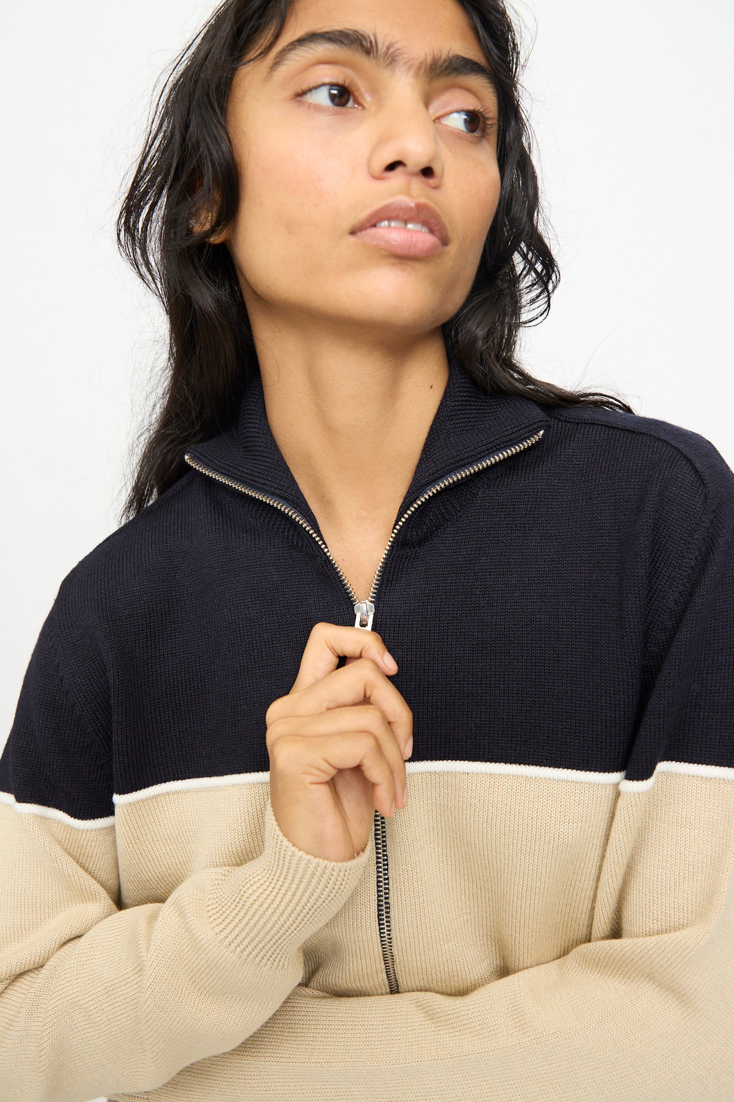 A person models the Cordera Merino Wool Cardigan in Colorblock, crafted in Spain with black and beige extrafine merino wool panels. They hold the zipper while looking to the side against a plain background.