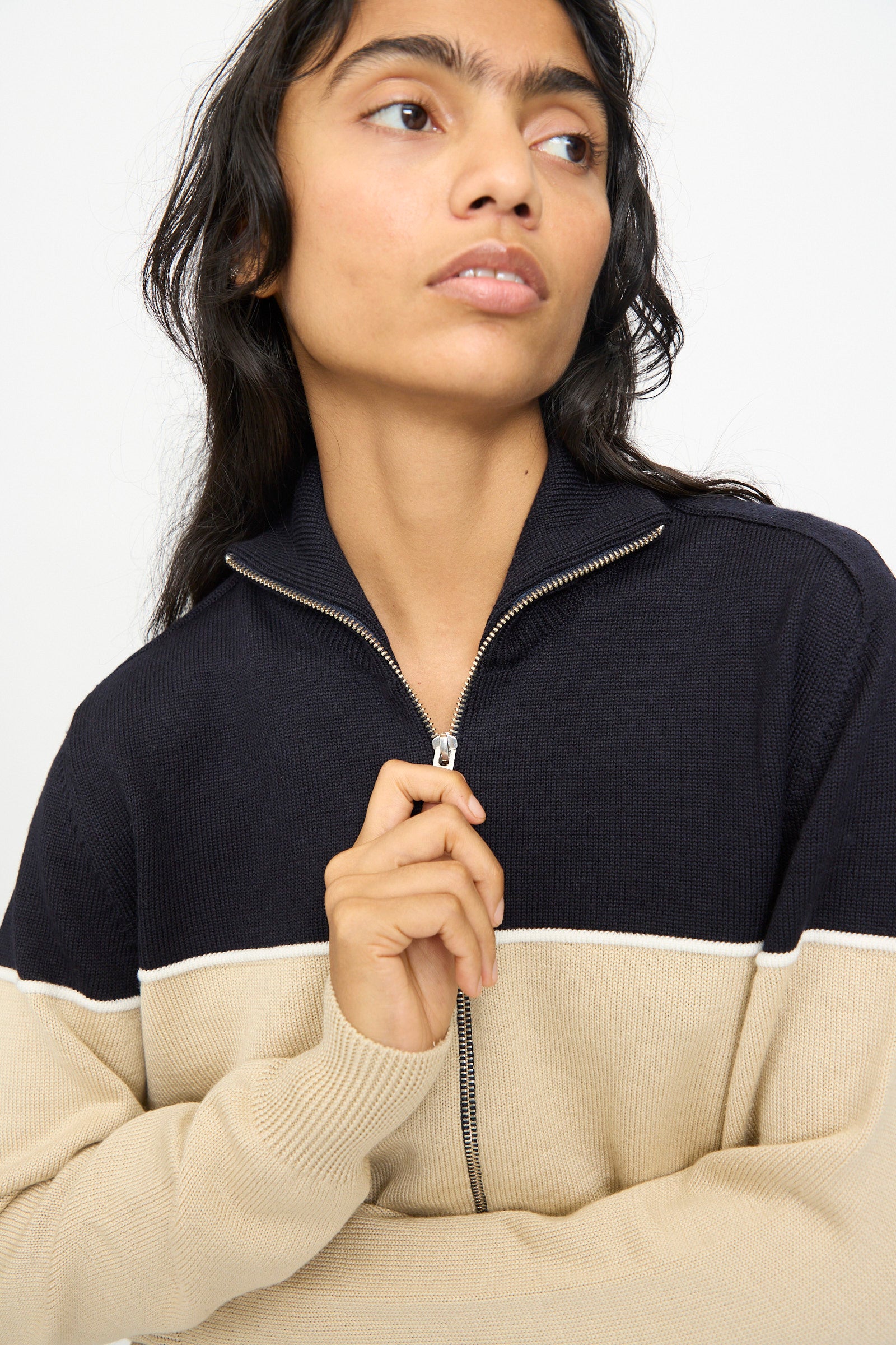A person models the Cordera Merino Wool Cardigan in Colorblock, crafted in Spain with black and beige extrafine merino wool panels. They hold the zipper while looking to the side against a plain background.