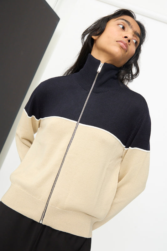 A person wearing the Cordera Merino Wool Cardigan in Colorblock, featuring a beige and black zip-up design crafted from extrafine merino wool with a high collar, stands indoors with hands behind their back and head tilted slightly upward.