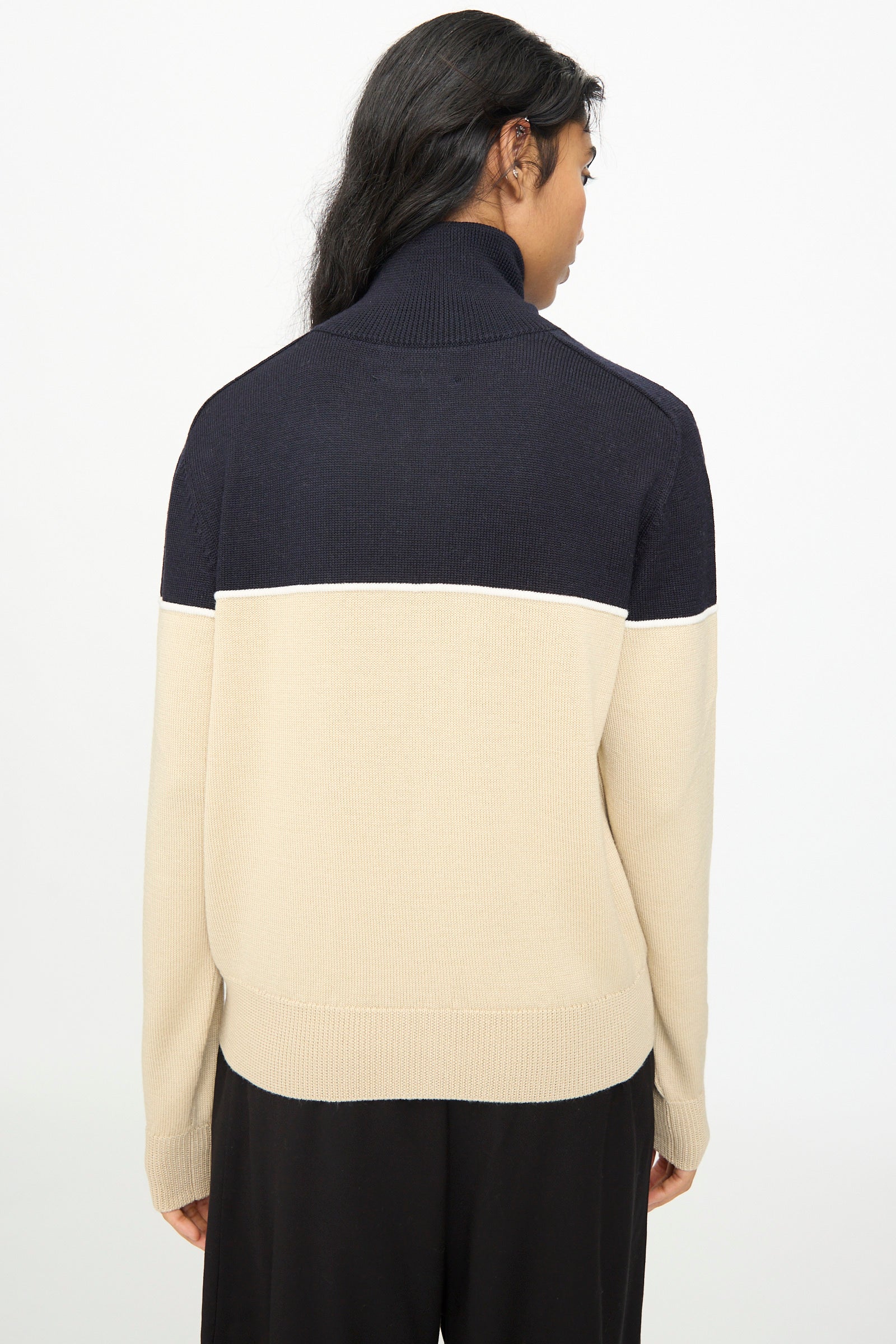 A person with long dark hair facing away wears the Cordera Merino Wool Cardigan in Colorblock—featuring beige and navy tones—paired with black pants, standing against a plain white background.