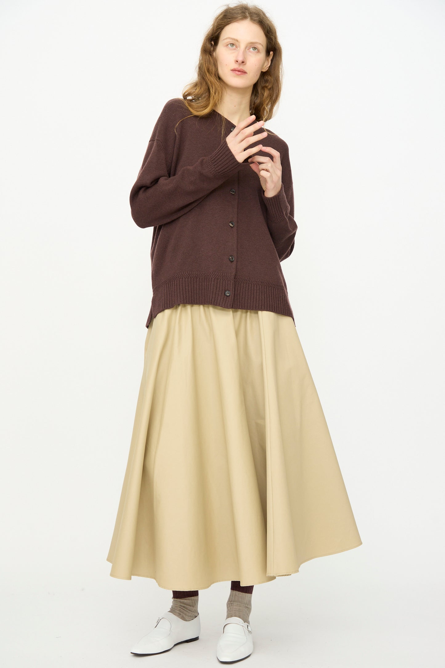 A person stands against a plain background wearing Cordera’s Merino Wool Cardigan in Prune, paired with a beige long skirt, brown socks, and white shoes.