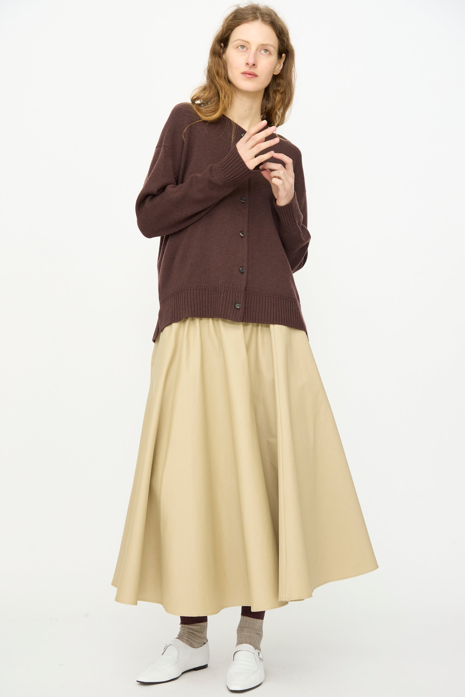 A person stands against a plain background wearing Cordera’s Merino Wool Cardigan in Prune, paired with a beige long skirt, brown socks, and white shoes.