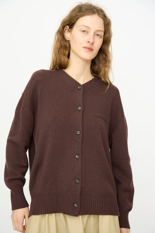 A person with long, wavy hair wears the Cordera Merino Wool Cardigan in Prune—made in Spain—over a light beige skirt, standing against a plain white background.