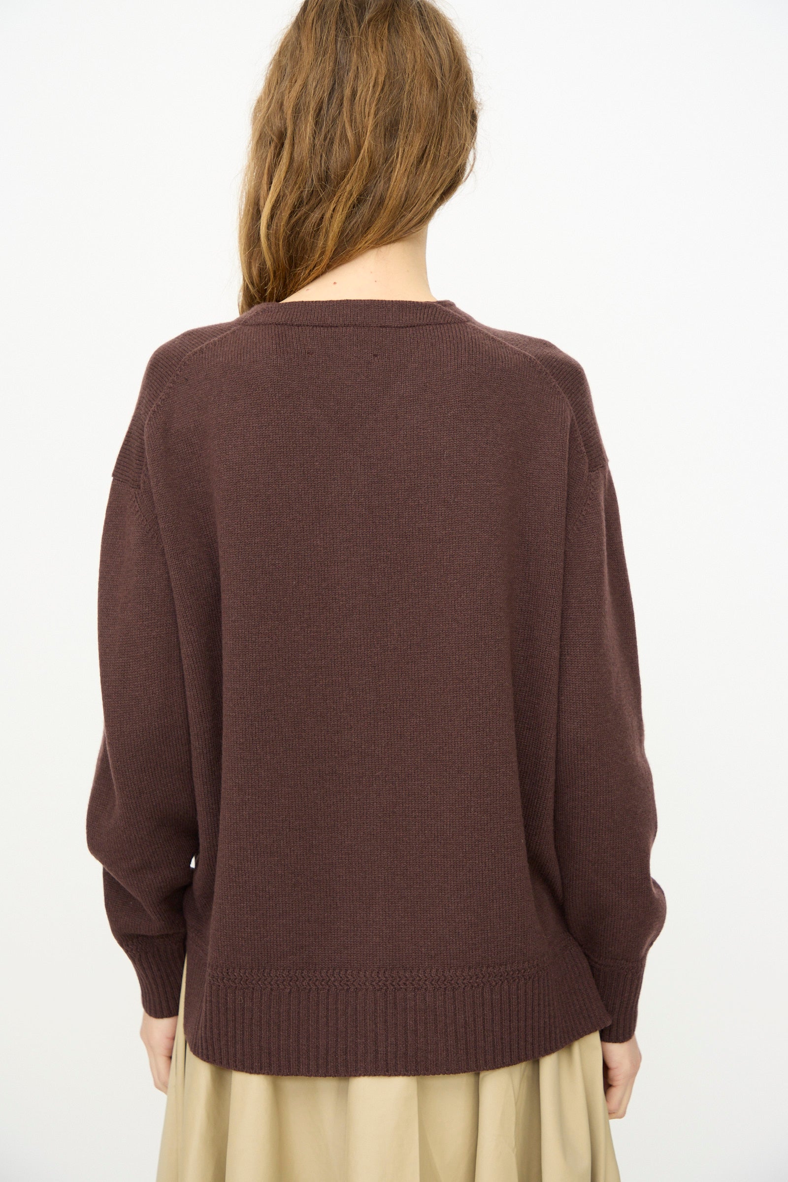 A person with long, wavy brown hair wears the Cordera Merino Wool Cardigan in Prune and a beige skirt, photographed from behind against a plain white background.
