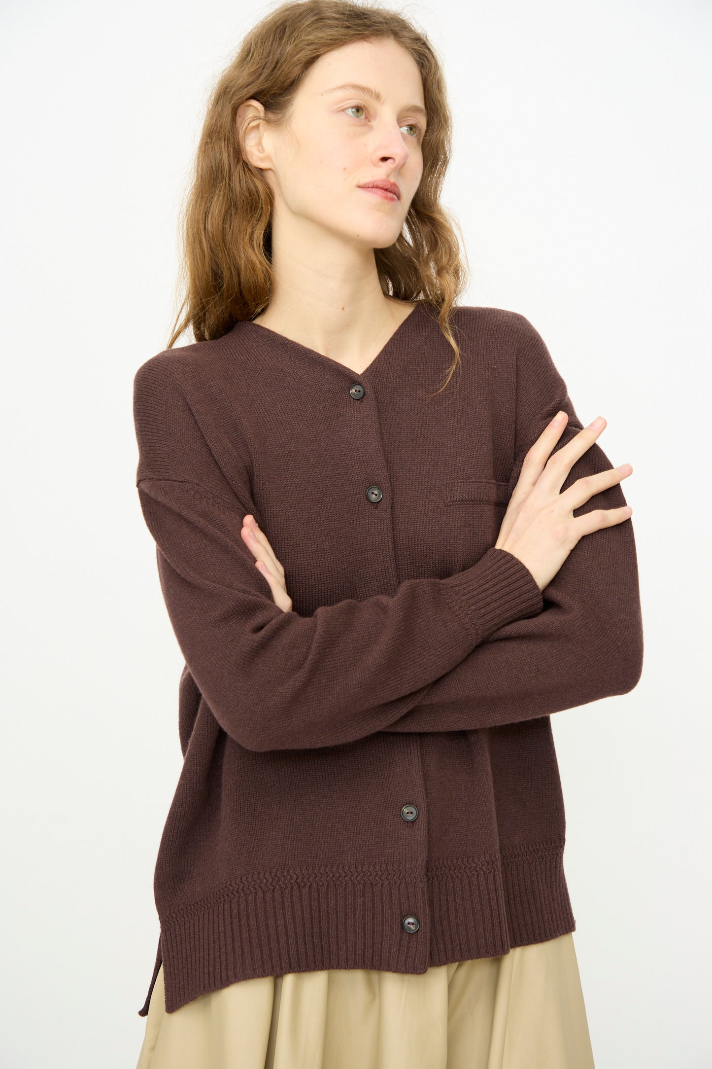 A woman with wavy brown hair wears Cordera’s mid-length Merino Wool Cardigan in Prune and a beige skirt, standing with arms crossed against a plain white background.