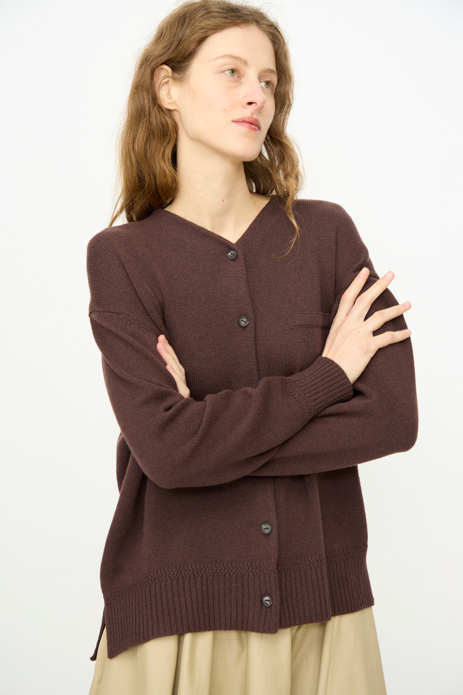 A woman with wavy brown hair wears Cordera’s mid-length Merino Wool Cardigan in Prune and a beige skirt, standing with arms crossed against a plain white background.