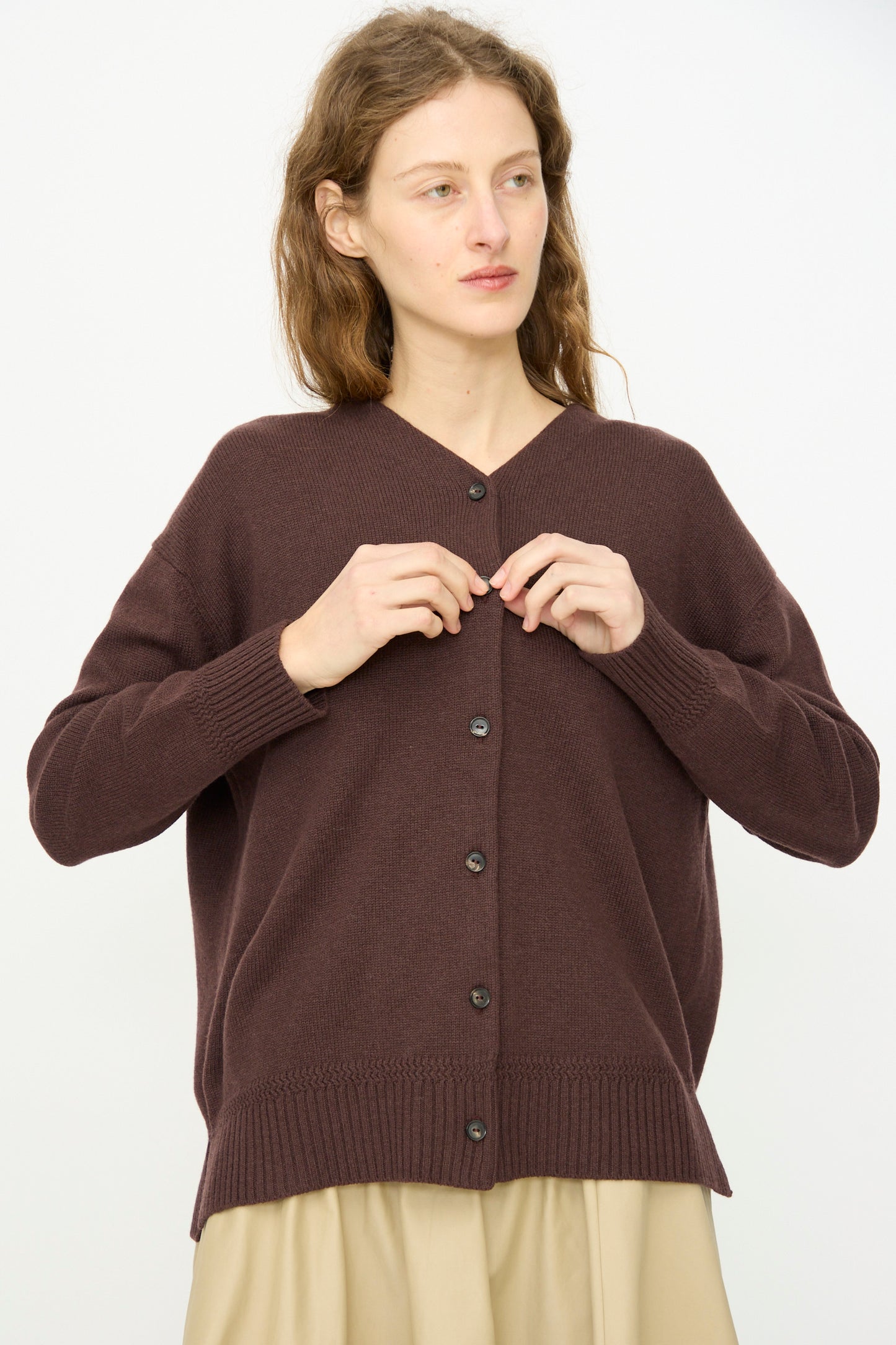 A woman with wavy hair wears Cordera’s Merino Wool Cardigan in Prune, a loose-fitting, mid-length ultrafine merino wool design, over a beige skirt while standing against a plain white background.