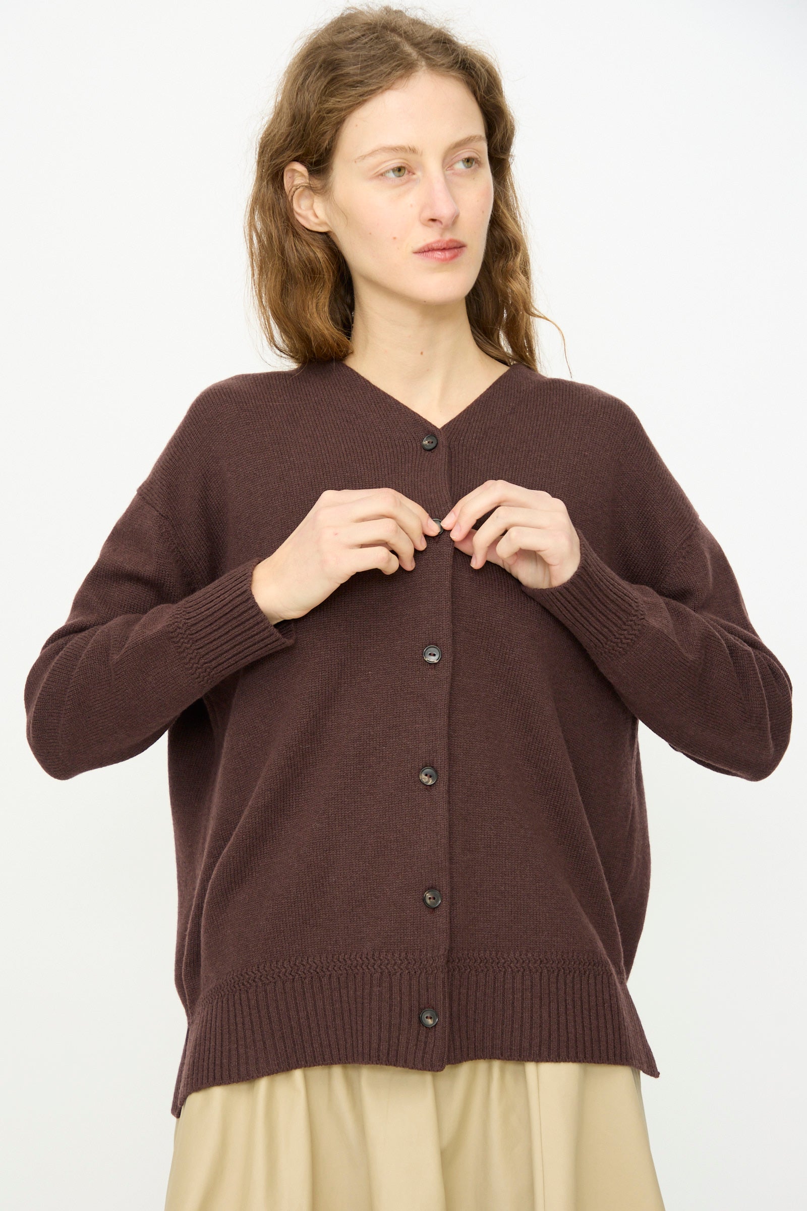 A woman with wavy hair wears Cordera’s Merino Wool Cardigan in Prune, a loose-fitting, mid-length ultrafine merino wool design, over a beige skirt while standing against a plain white background.