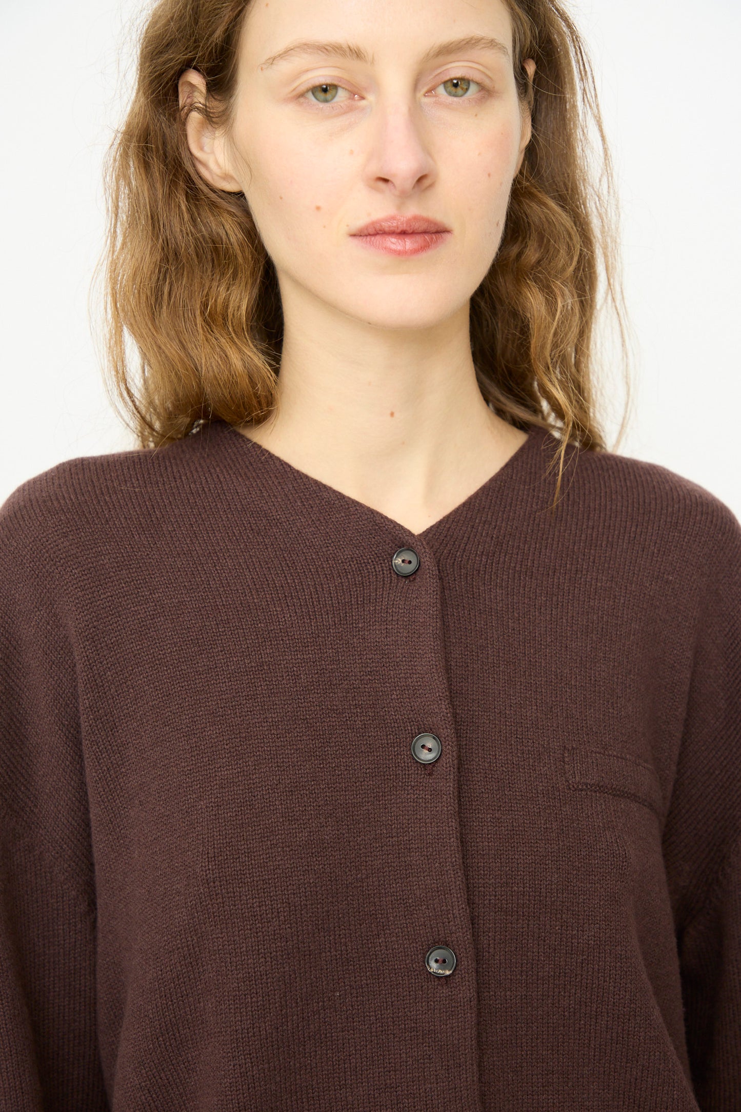 A woman with wavy brown hair wears Cordera's Merino Wool Cardigan in Prune, a dark brown ultrafine merino wool mid-length piece, standing against a plain white background.