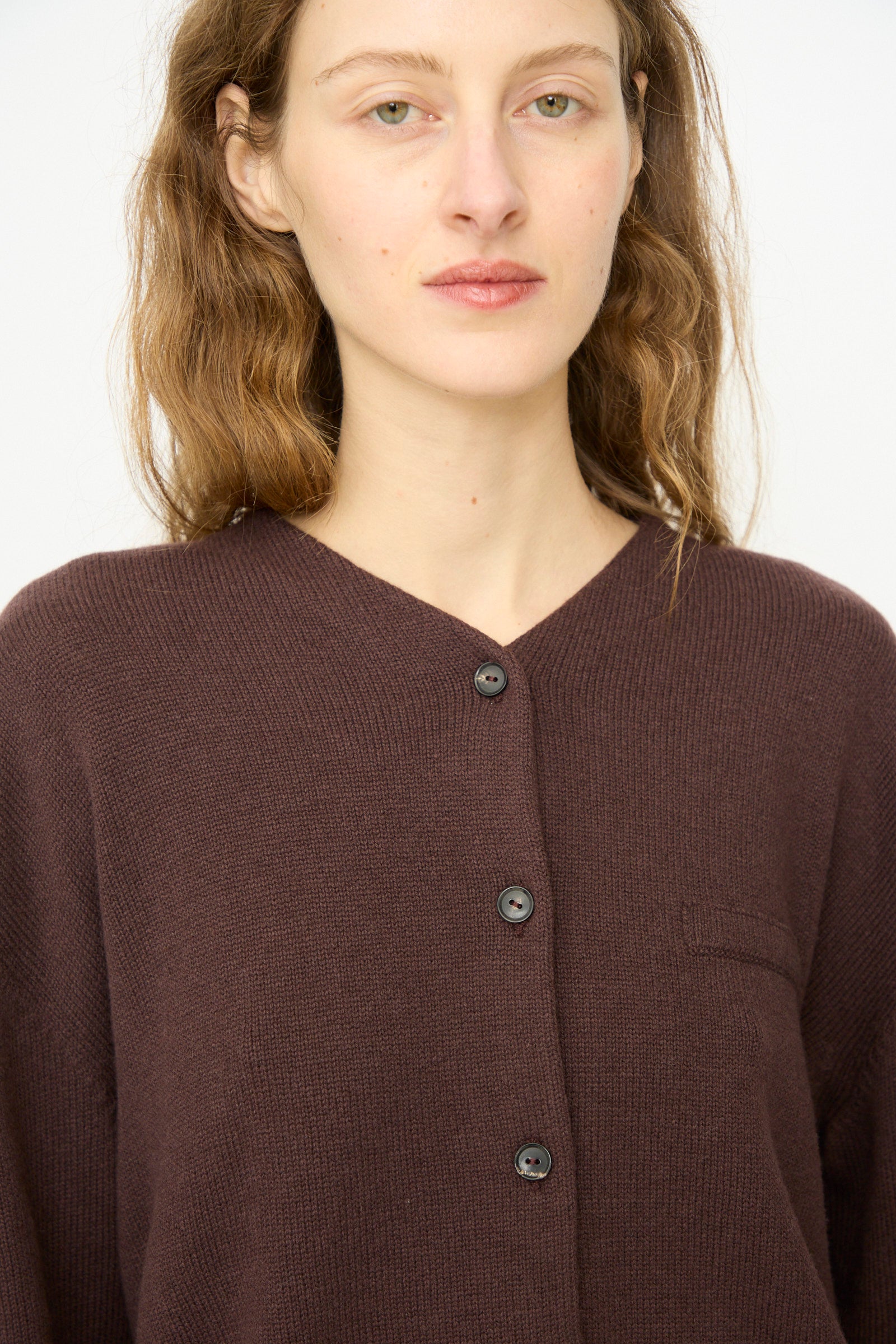 A woman with wavy brown hair wears Cordera's Merino Wool Cardigan in Prune, a dark brown ultrafine merino wool mid-length piece, standing against a plain white background.
