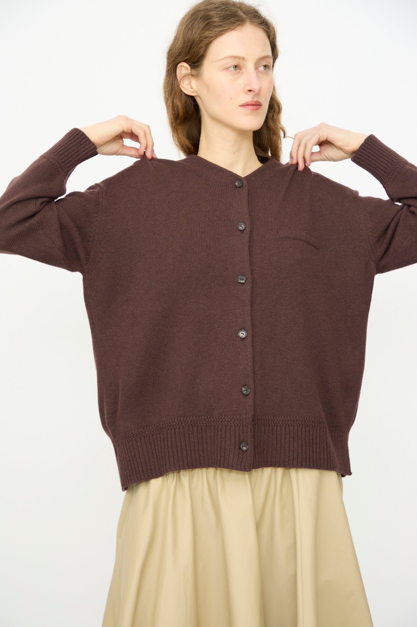 A person wearing Cordera's loose, mid-length Merino Wool Cardigan in Prune and a beige skirt stands against a plain white background.