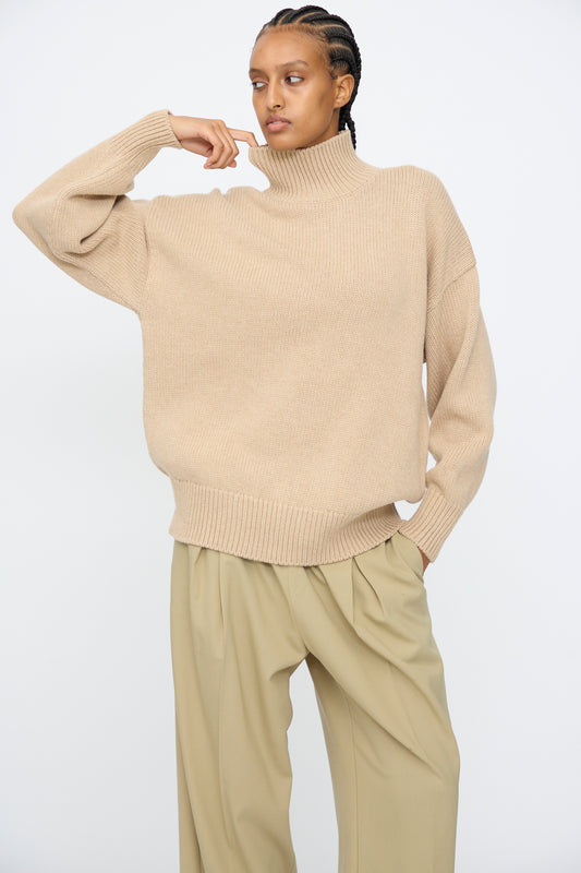 A person wearing the Cordera Merino Wool Chunky Turtleneck Sweater in Beige and wide-leg khaki pants stands against a plain white background, one hand raised near their face.
