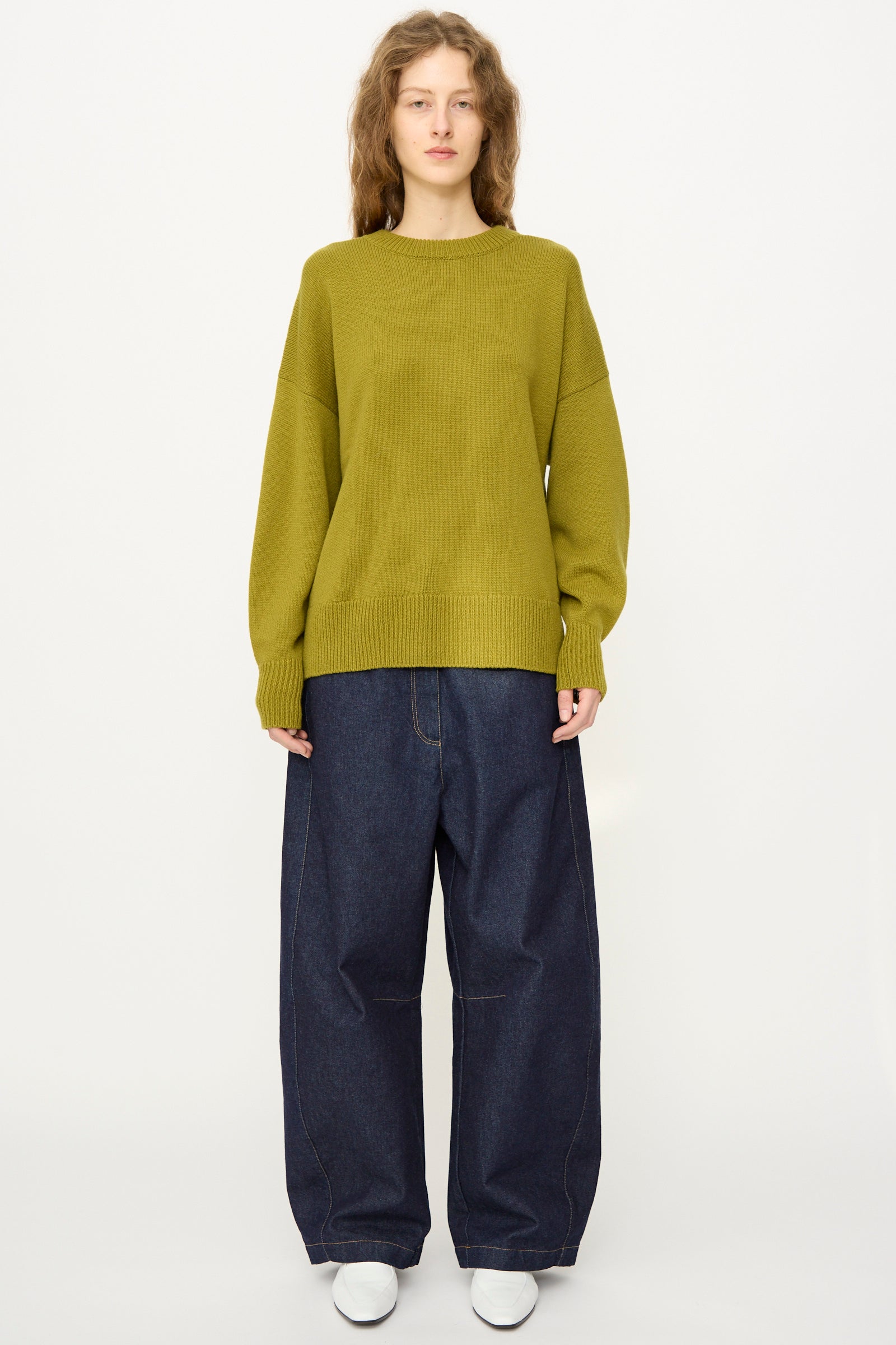 A person stands against a plain background wearing Cordera’s Merino Wool Oversized Sweater in Woodbine, wide-leg dark blue jeans, and white shoes. The sweater is made in Spain. They face forward with a neutral expression.