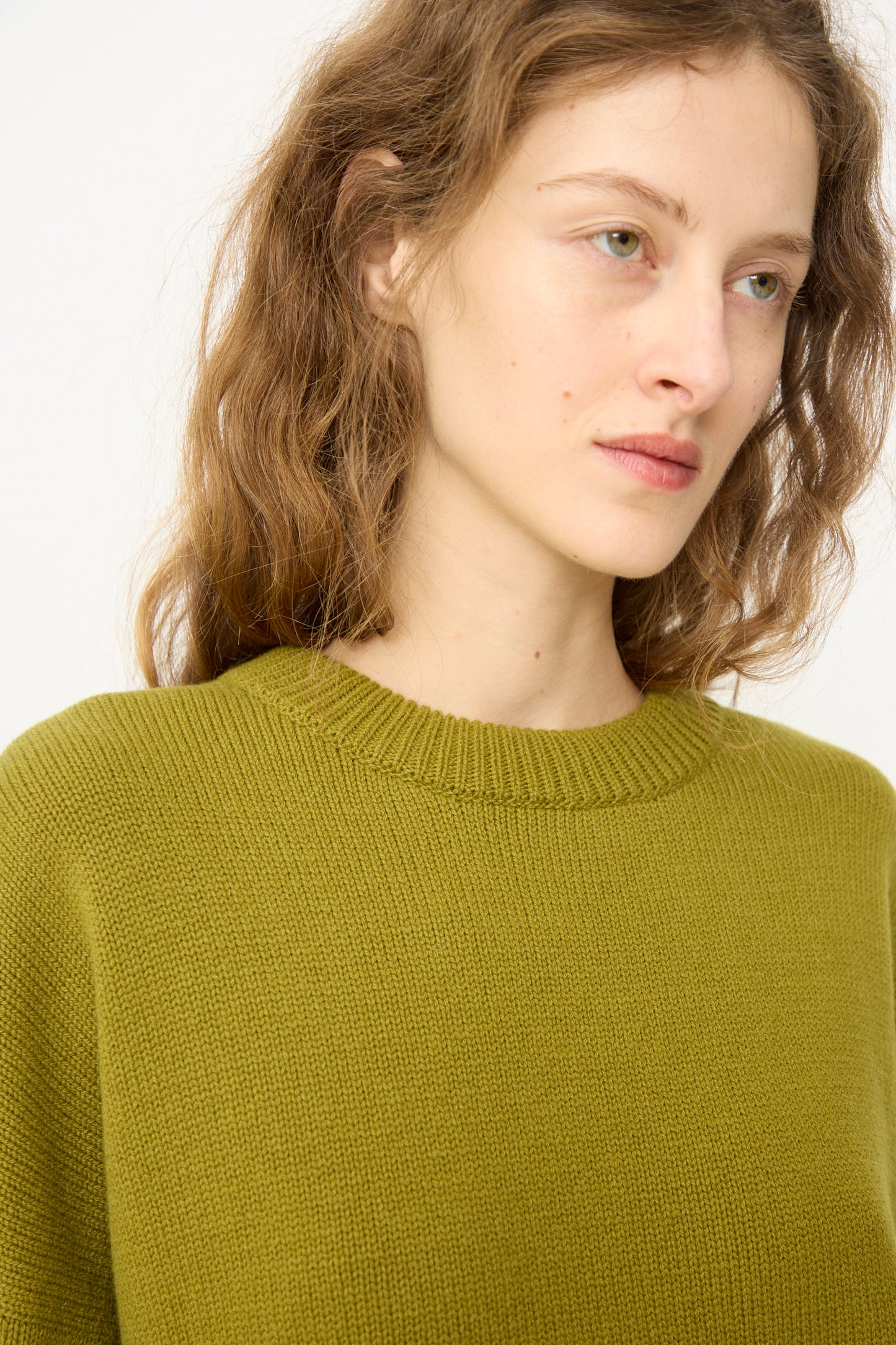A person with wavy light brown hair models the Cordera Merino Wool Oversized Sweater in Woodbine, glancing to the side against a plain background.