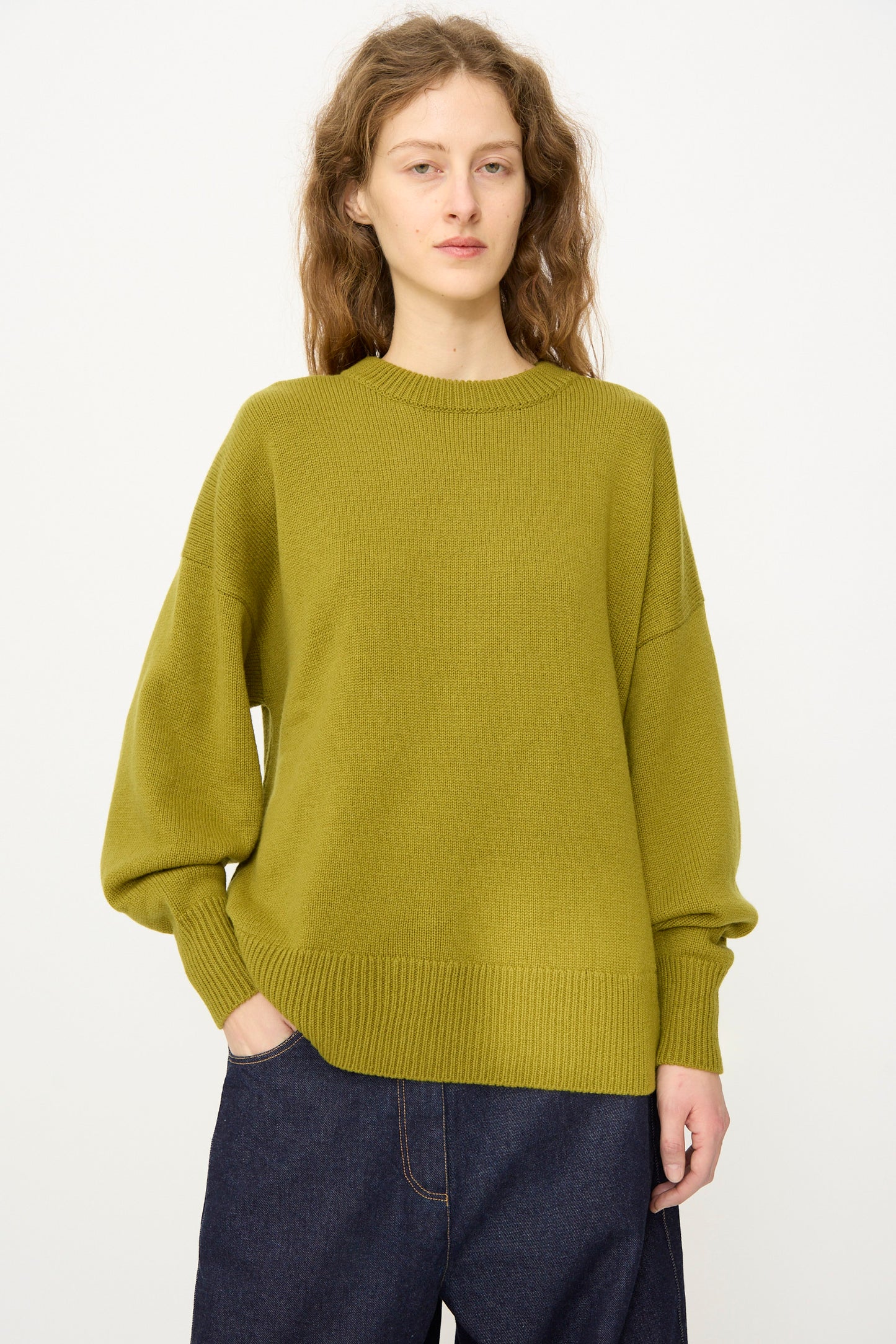 A person with long wavy hair wears the Cordera Merino Wool Oversized Sweater in Woodbine and dark jeans, standing against a plain light background with one hand in their pocket.