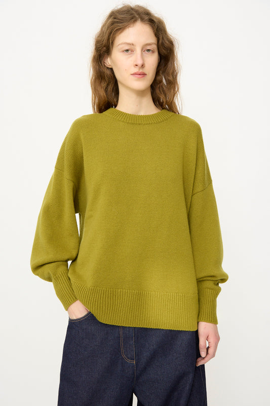 A person with long wavy hair wears the Cordera Merino Wool Oversized Sweater in Woodbine and dark jeans, standing against a plain light background with one hand in their pocket.