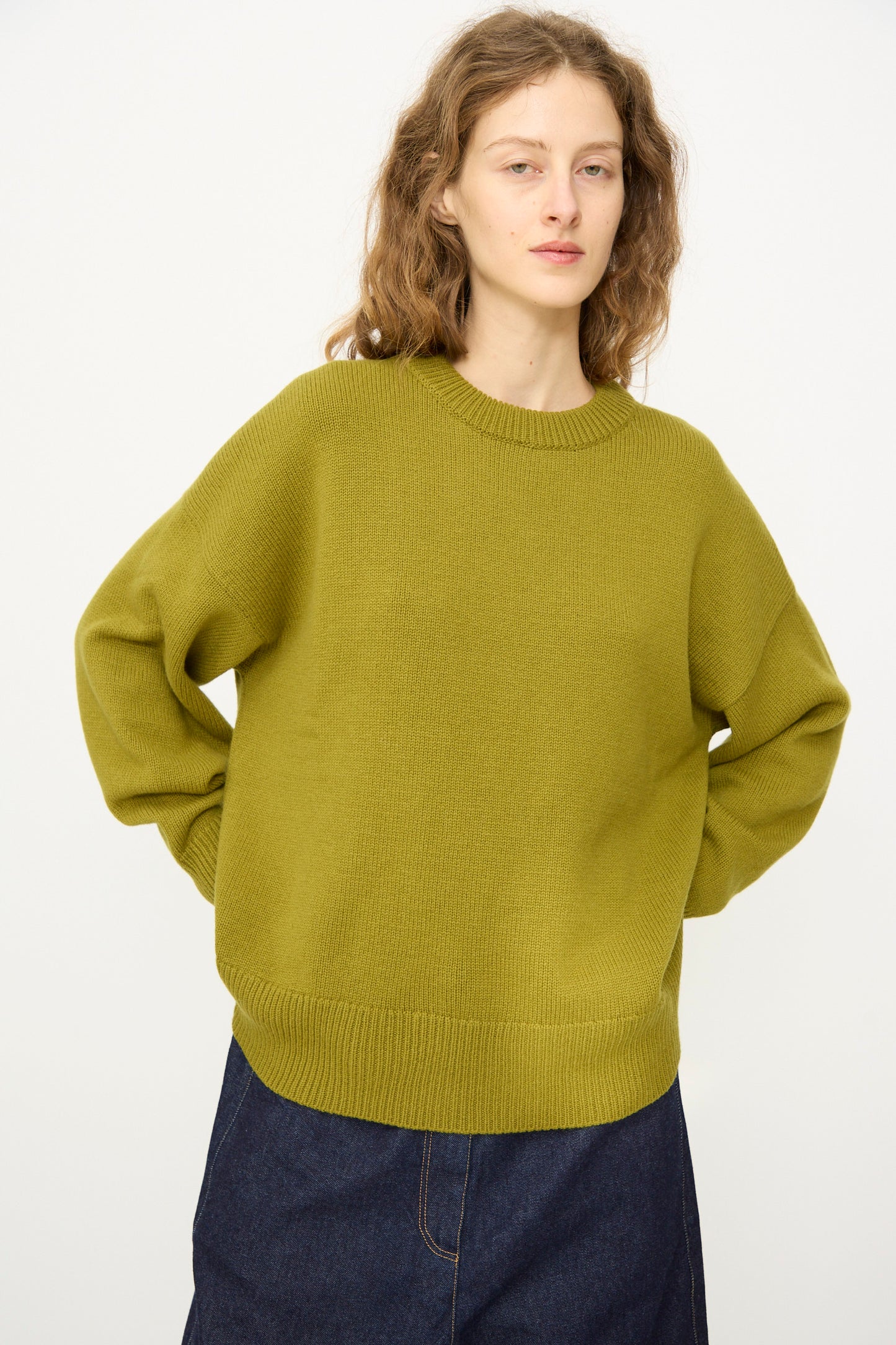 A person wears the Cordera Merino Wool Oversized Sweater in Woodbine and a dark denim skirt, standing against a plain white background.