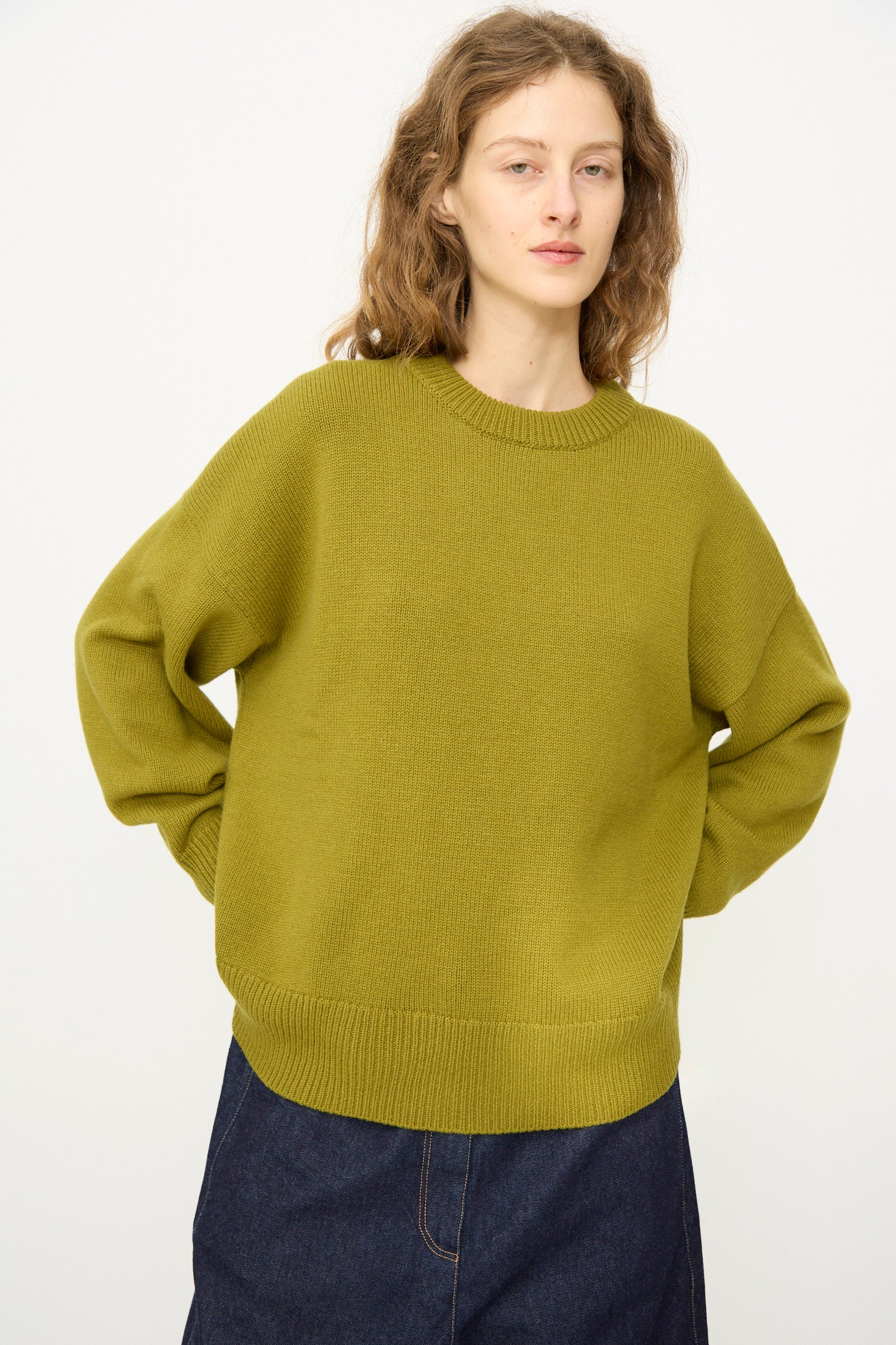 A person wears the Cordera Merino Wool Oversized Sweater in Woodbine and a dark denim skirt, standing against a plain white background.
