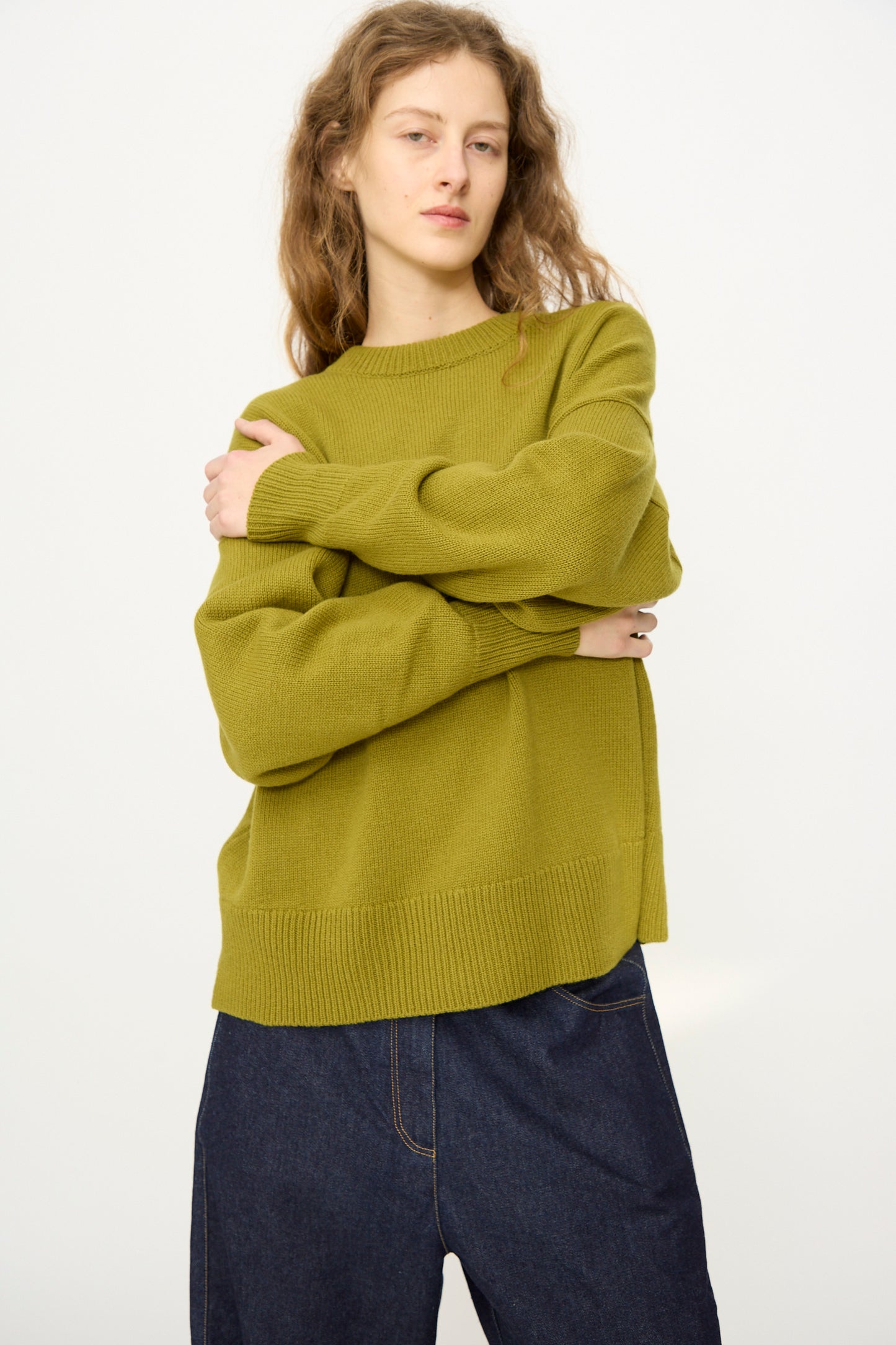 A person with long, wavy hair wears the Cordera Merino Wool Oversized Sweater in Woodbine and loose dark blue jeans, standing against a plain white background.