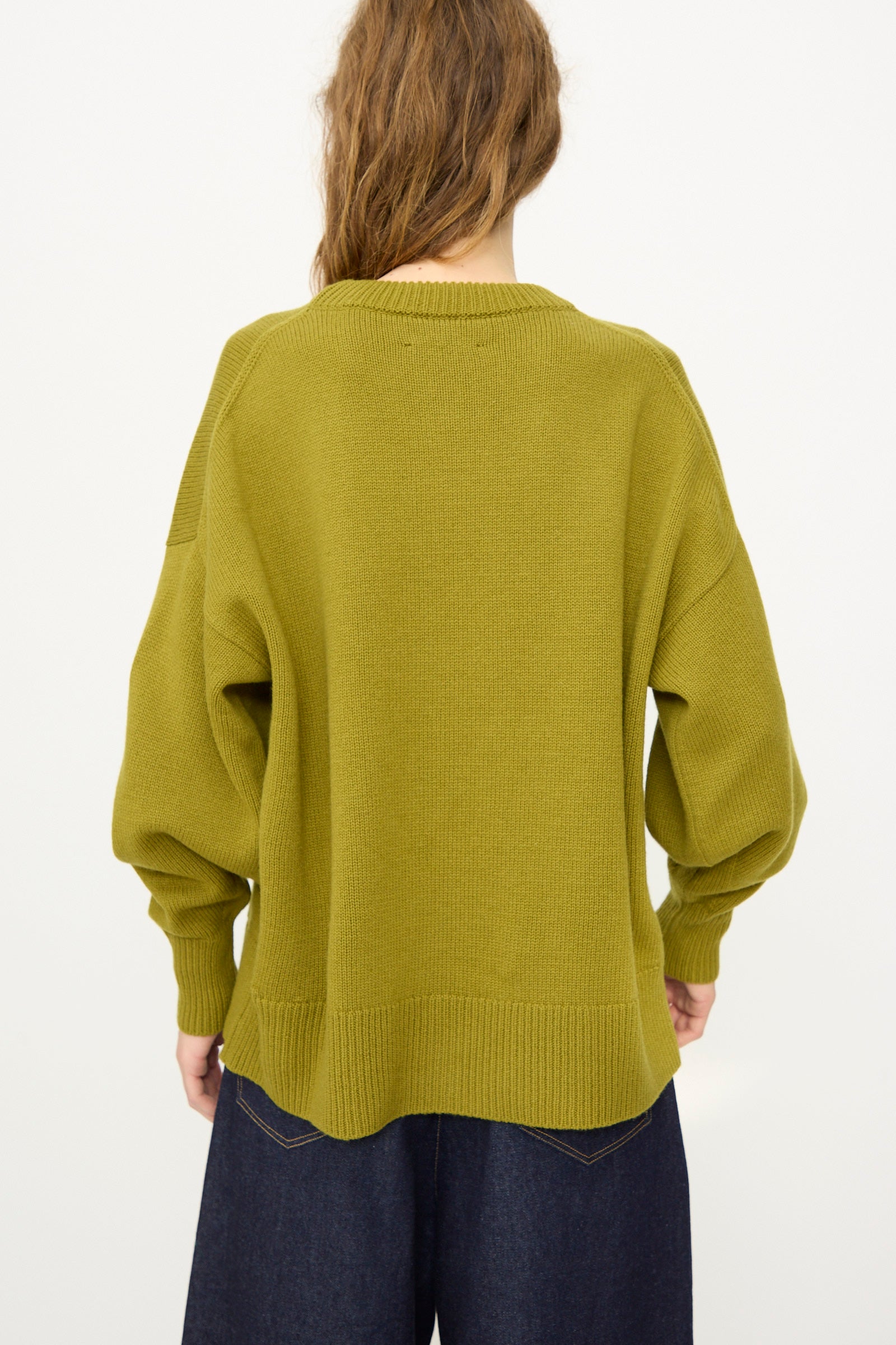 Model wearing the Cordera Merino Wool Oversized Sweater in Woodbine, featuring long sleeves and an olive green hue, paired with dark pants and shown from the back against a plain white background.