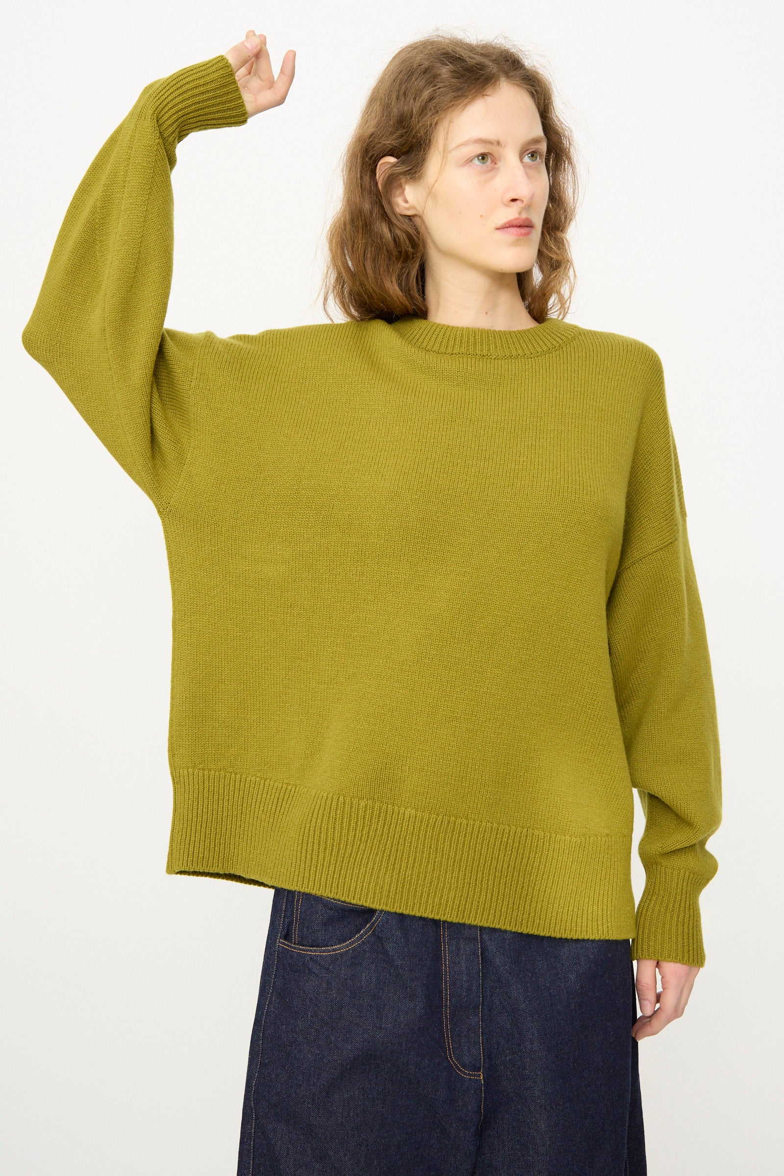 A woman wears the Cordera Merino Wool Oversized Sweater in Woodbine with a dark denim skirt, standing with one arm raised against a plain background.