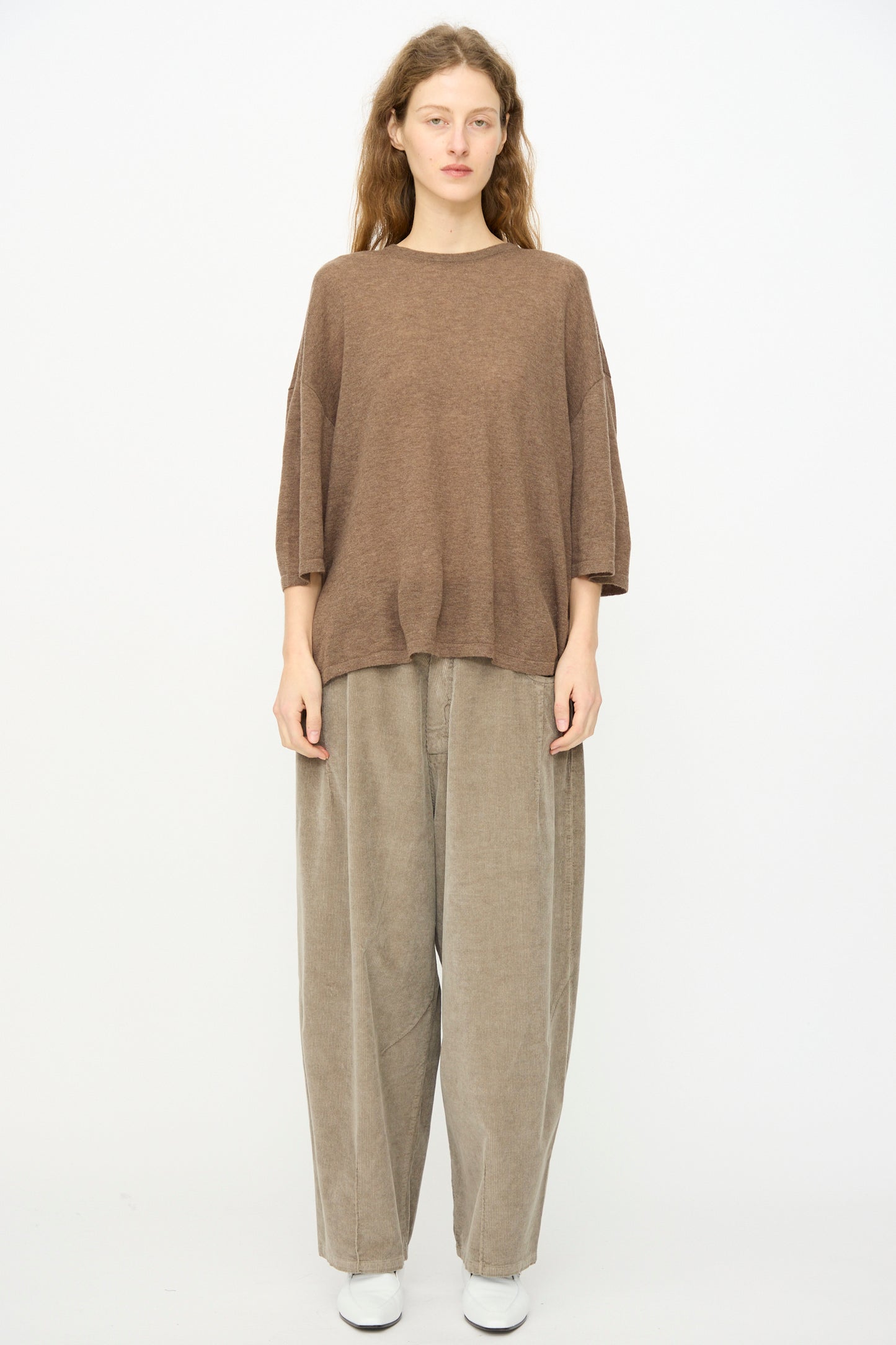 A person stands against a plain background, facing forward with a neutral expression, wearing Cordera's Merino Wool Oversized T-Shirt in Deep Taupe, paired with wide beige pants and white shoes.