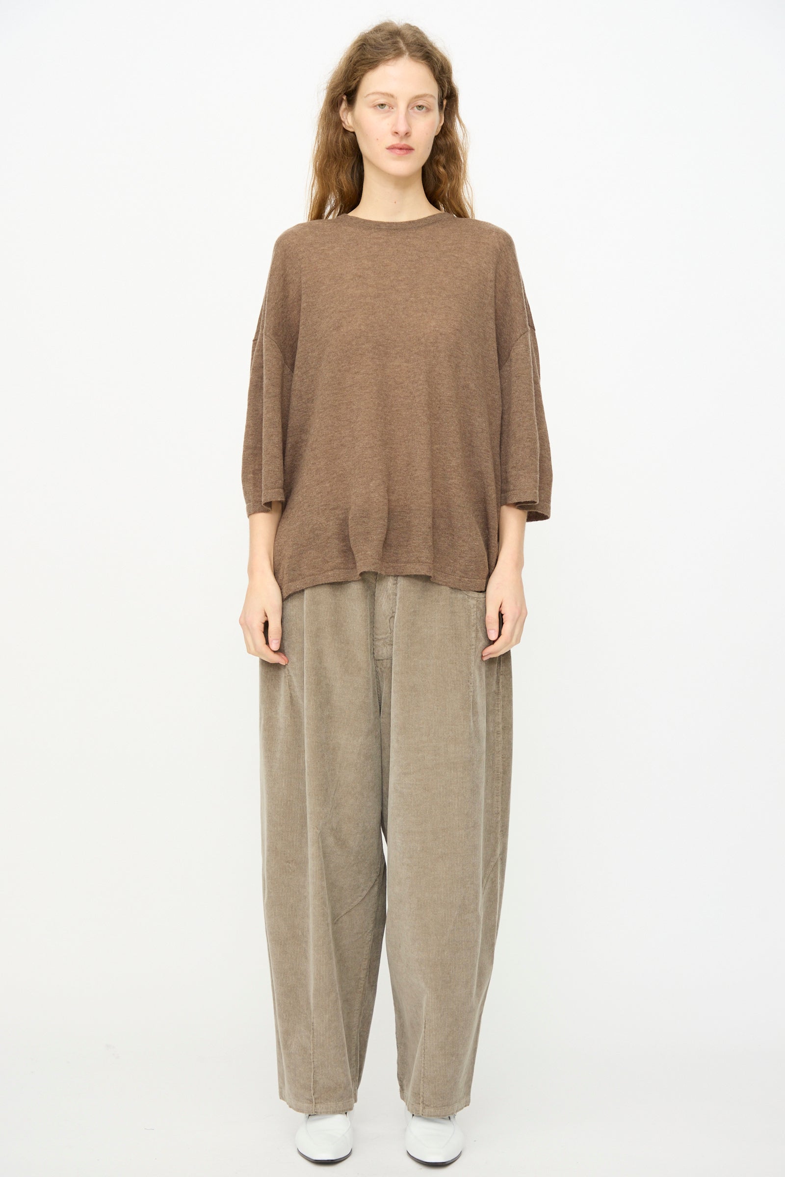 A person stands against a plain background, facing forward with a neutral expression, wearing Cordera's Merino Wool Oversized T-Shirt in Deep Taupe, paired with wide beige pants and white shoes.