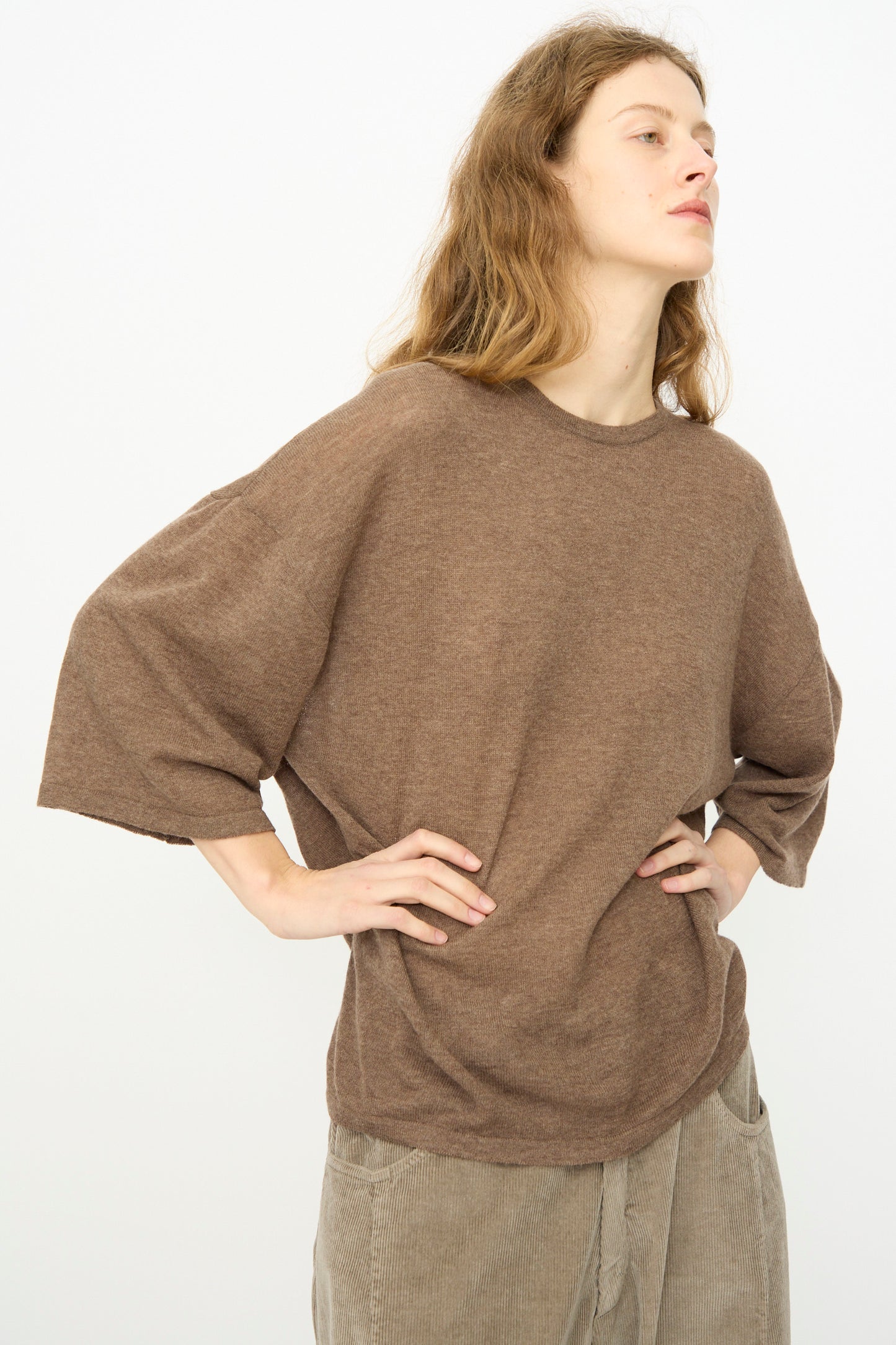 A person with long hair wears Cordera's Merino Wool Oversized T-Shirt in Deep Taupe and light brown pants, standing with hands on hips against a plain light background.