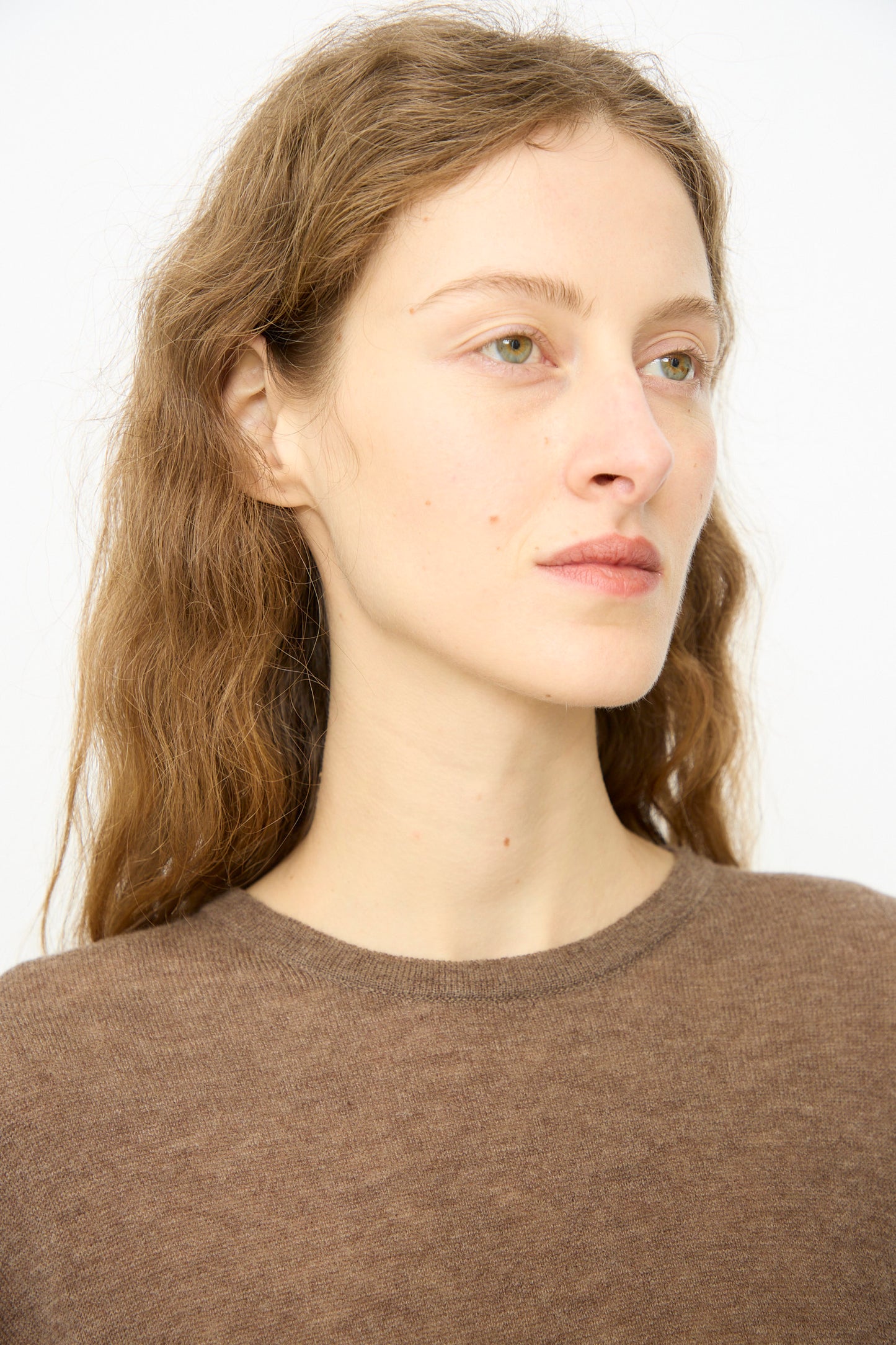 A person with long, wavy brown hair and light skin wears the Cordera Merino Wool Oversized T-Shirt in Deep Taupe, gazing slightly to the side against a plain light background.